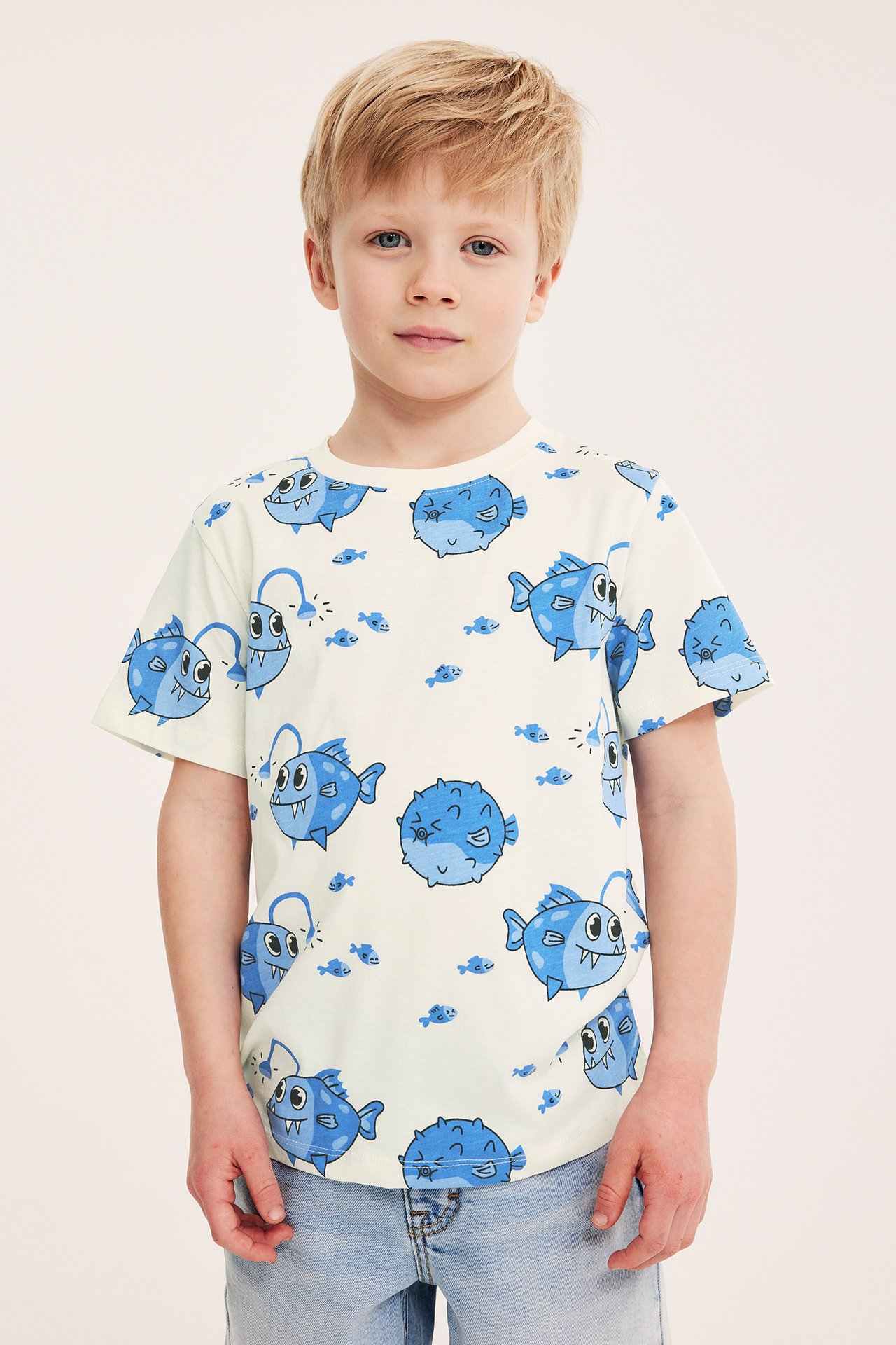 Frontvy av ett barn i en offwhite bomulls-T-shirt med blått fiskmönster och ljusblå jeans.