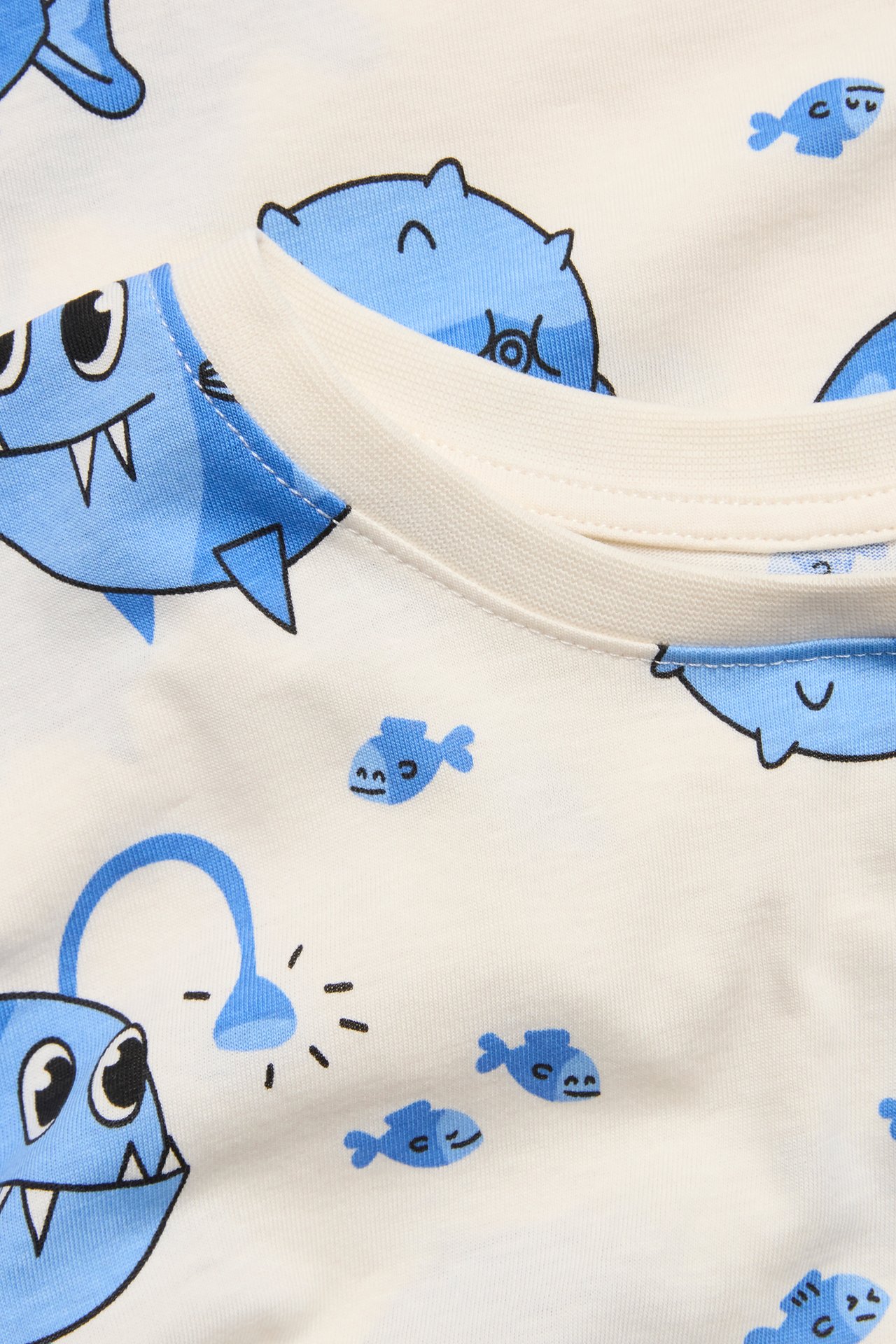 Närbild på en offwhite t-shirt i bomull för barn med blått tecknat fiskmönster och ribbad halsringning.