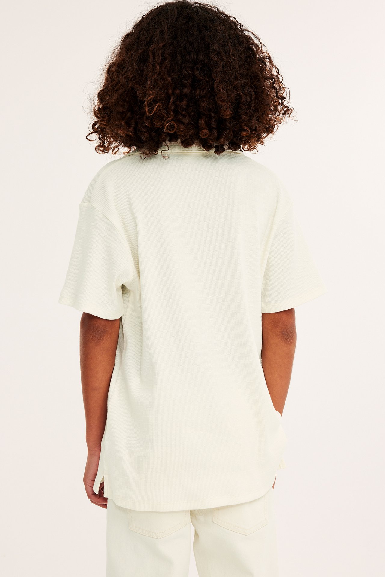 Widok z tyłu dziecka w kremowej, strukturalnej koszulce polo z krótkim rękawem i kremowych spodniach.