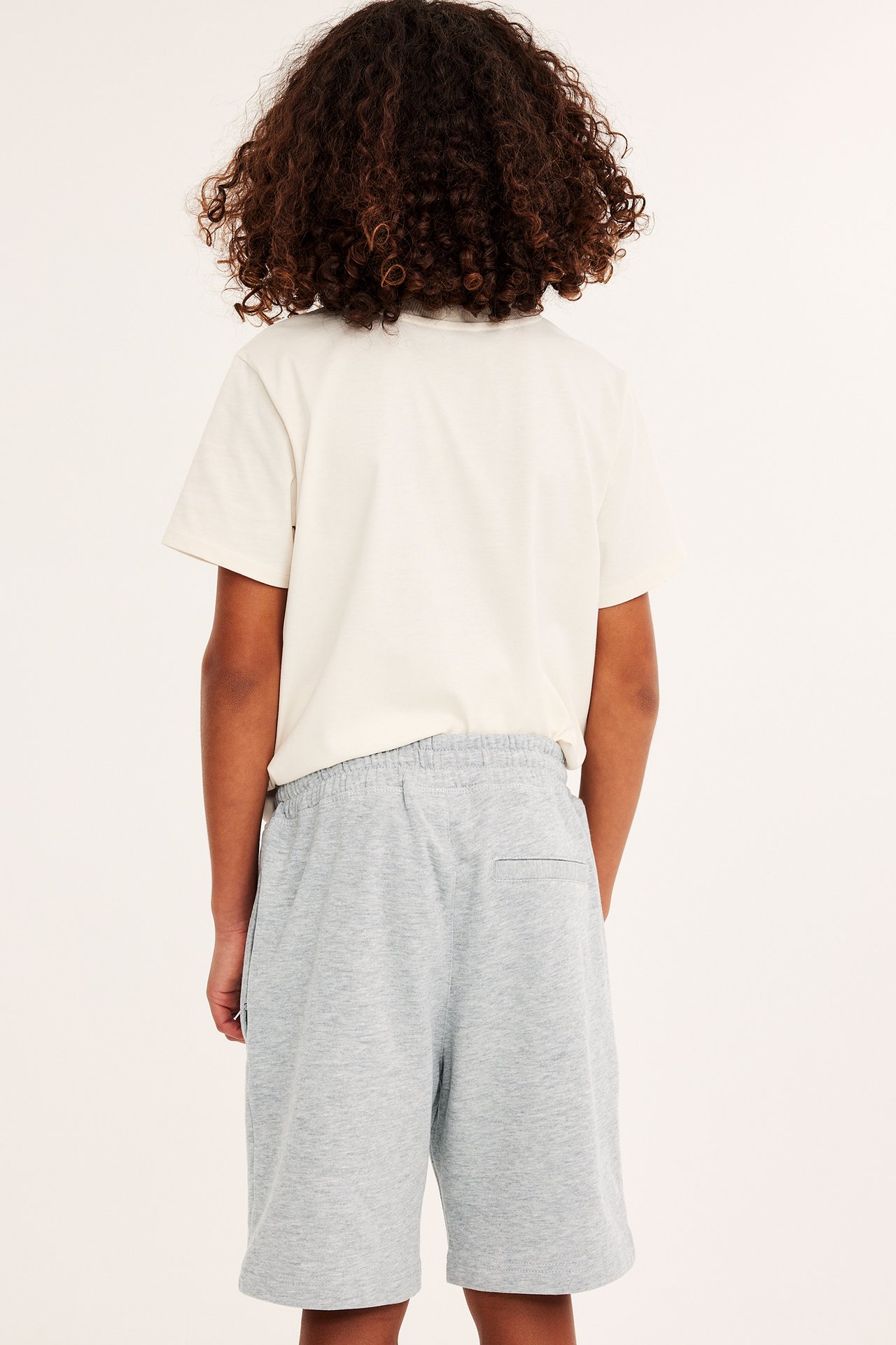 Bakifrån, ett barn i ljusgrå mjukisshorts med resår i midjan och en ljusbeige t-shirt.