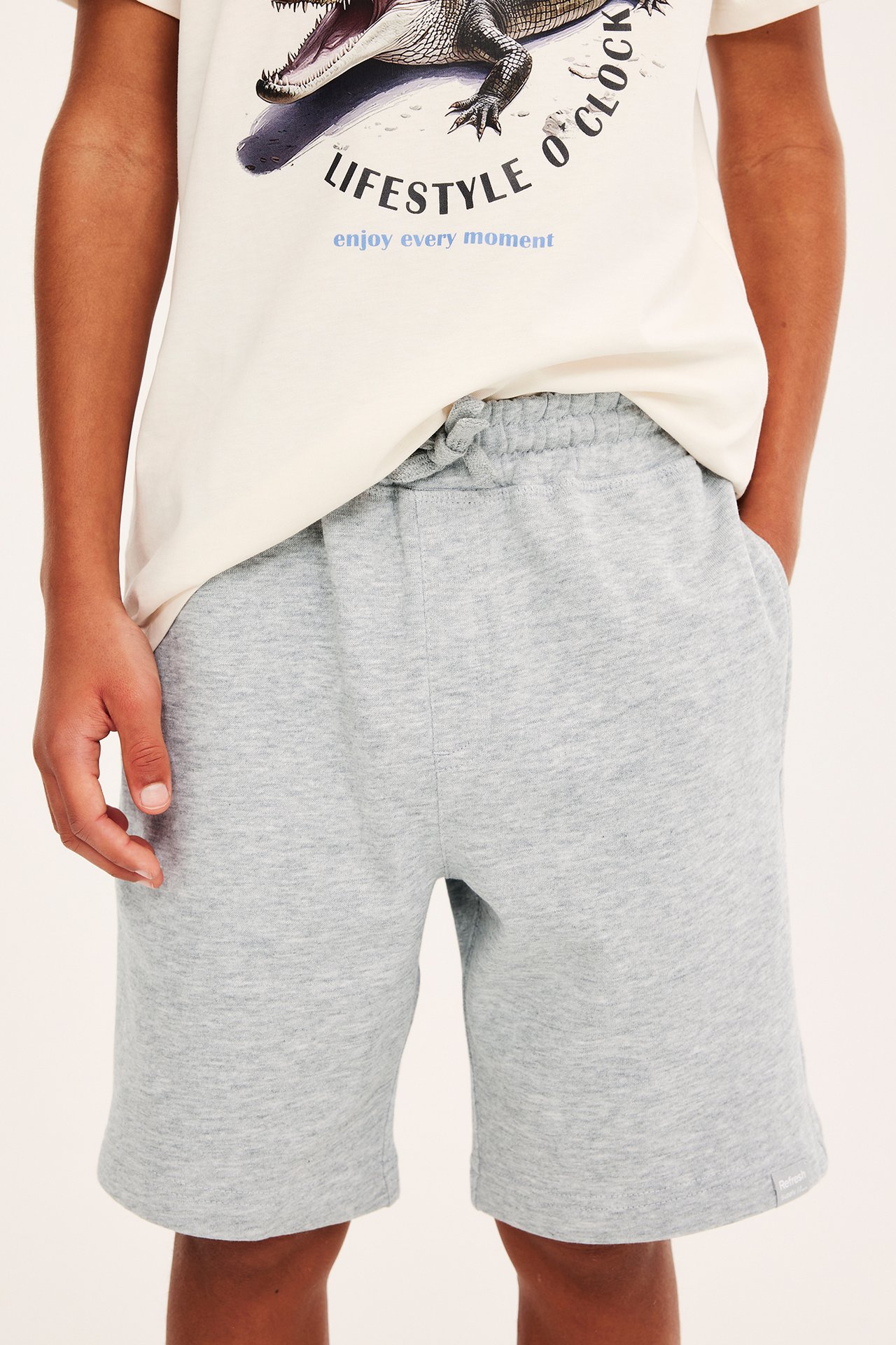 Frontvy av ett barn i ljusgrå mjukisshorts med dragsko och en krämfärgad t-shirt med tryck.