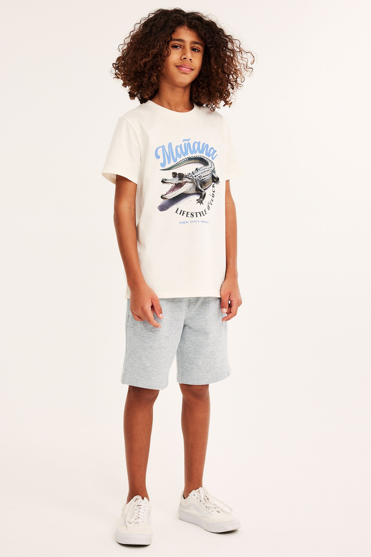 Frontvy: Barn i ljusgrå mjukisshorts i bomullsblandning med dragsko, gräddvit t-shirt med krokodilmönster, vita sneakers.