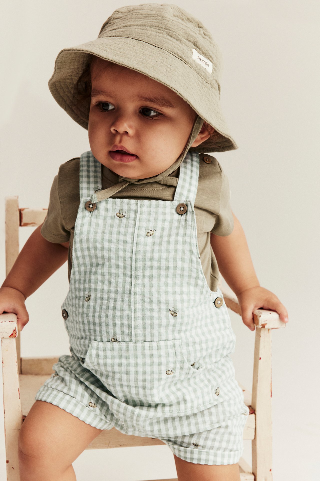 Frontvisning: Baby i lysegrønne rutete snekkerbukser med biebroderi, khaki t-skjorte og beige bøttehatt.