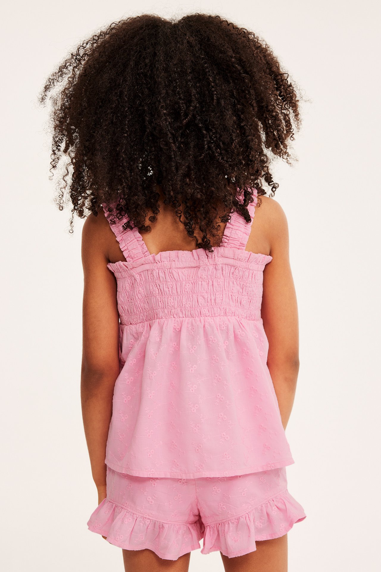 Bakifrån: ett barn i rosa broderad bomullstopp med smock och matchande volangshorts.