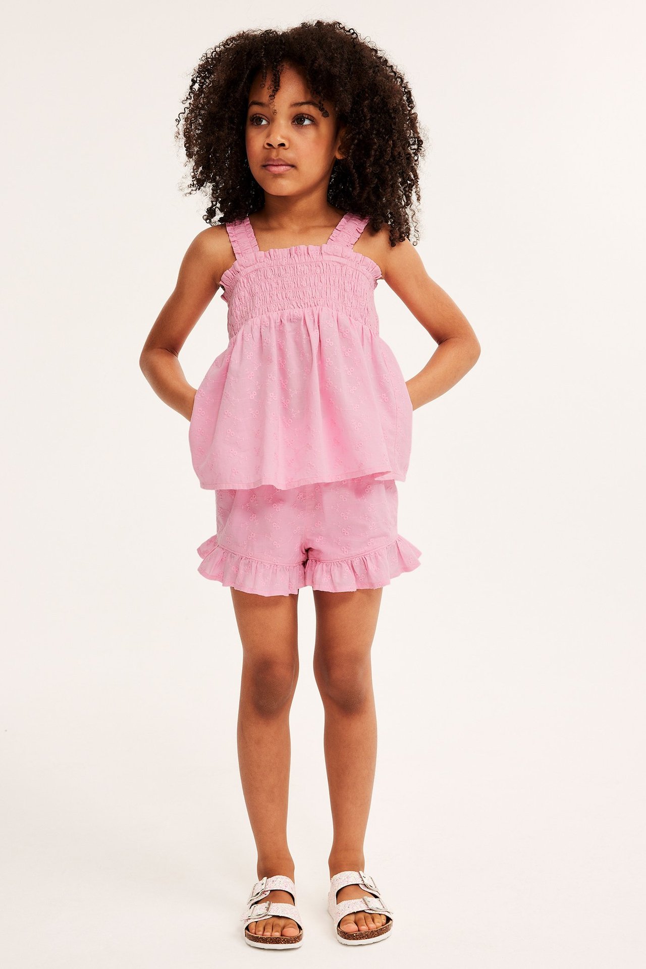 Framifrån: ett barn i rosa broderad bomullstopp med smock, matchande volangshorts och vita sandaler.