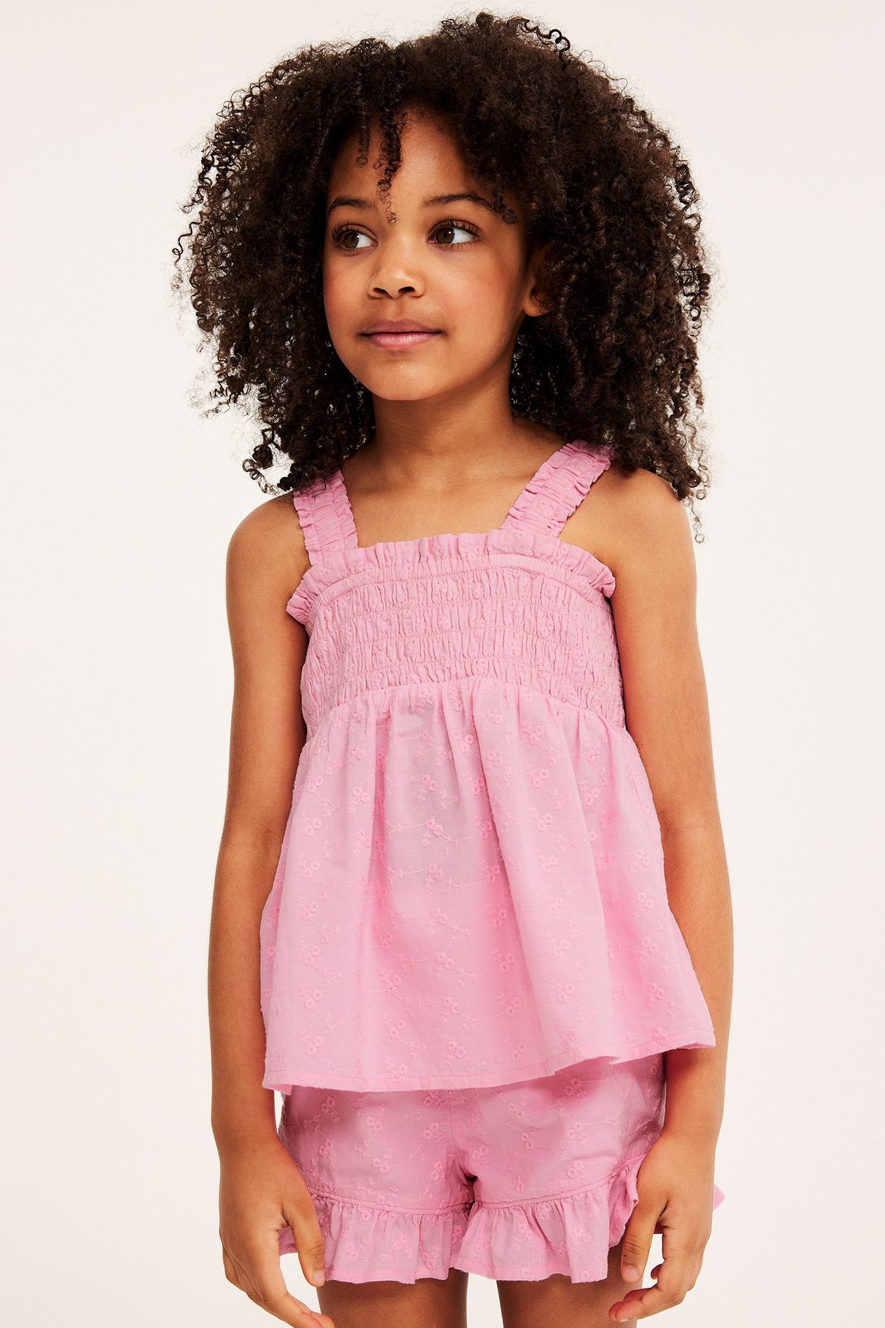 Framifrån, ett barn i rosa broderad bomullstopp med smock och matchande volangshorts.