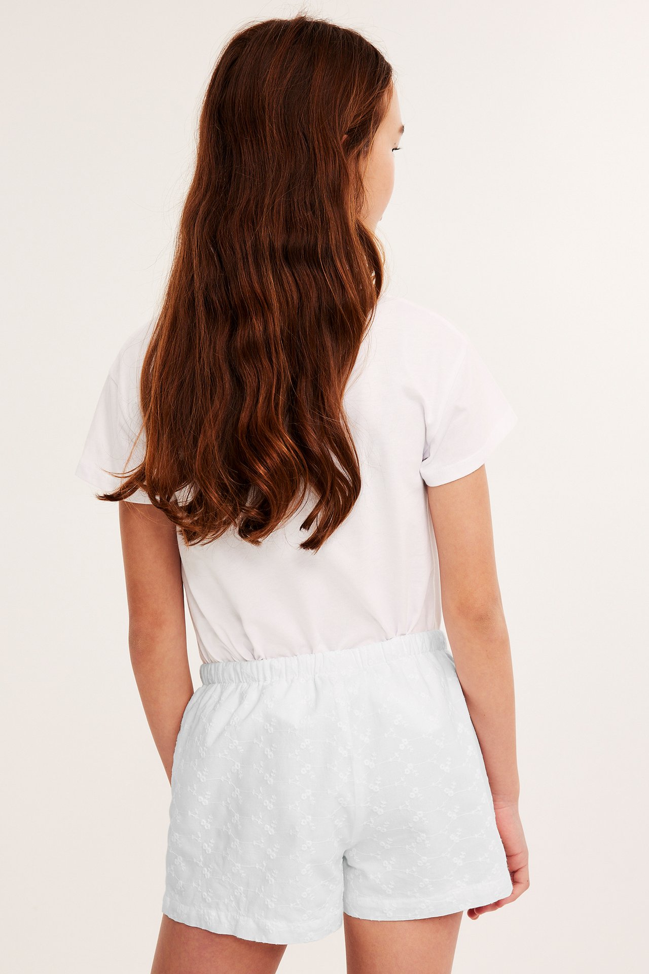 Bakifrån: Kid bär vita broderade bomullsshorts med resår i midjan och en vit t-shirt.