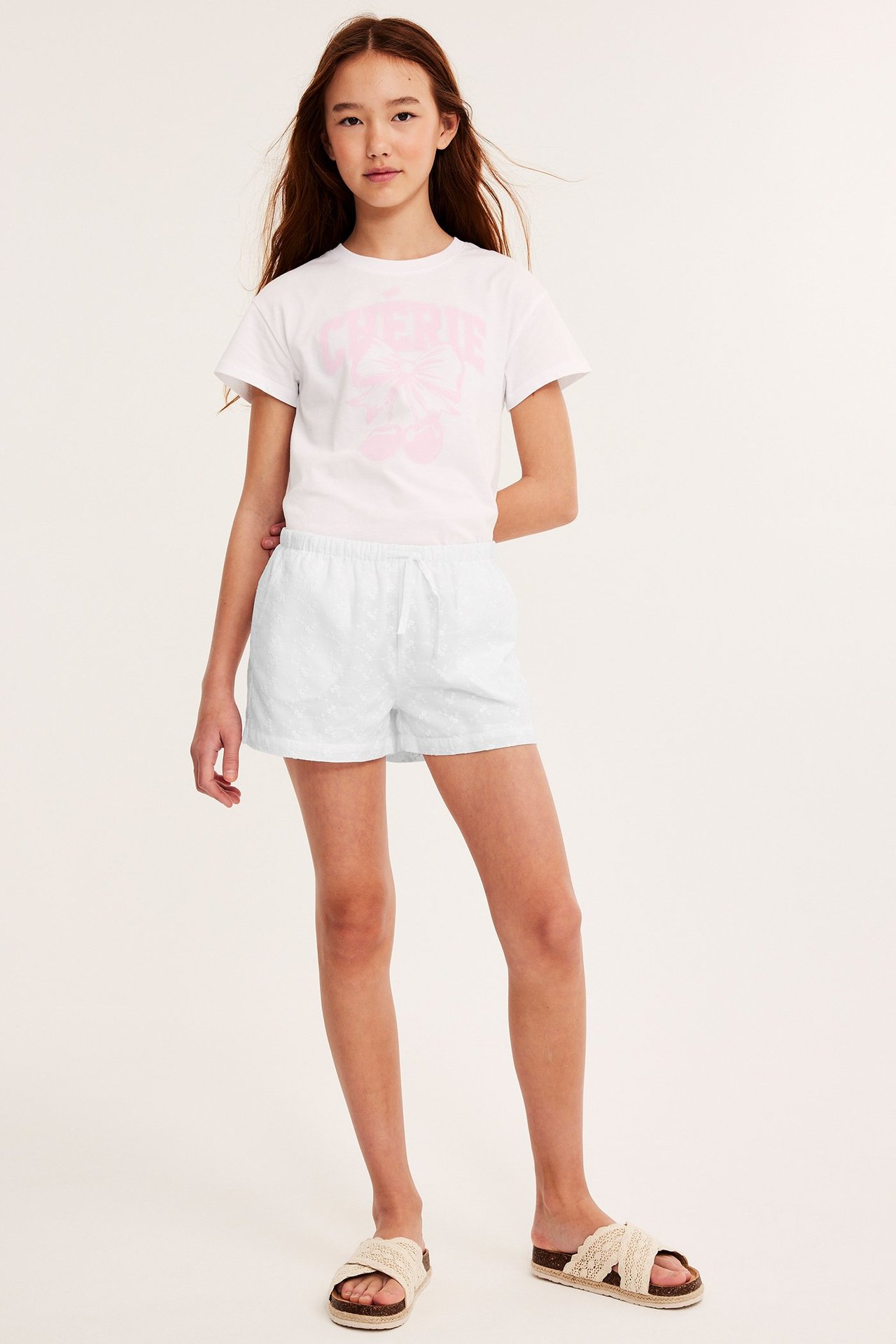 Helbild framifrån på ett barn i vita broderade bomullsshorts, en vit t-shirt med rosa tryck och beiga sandaler.