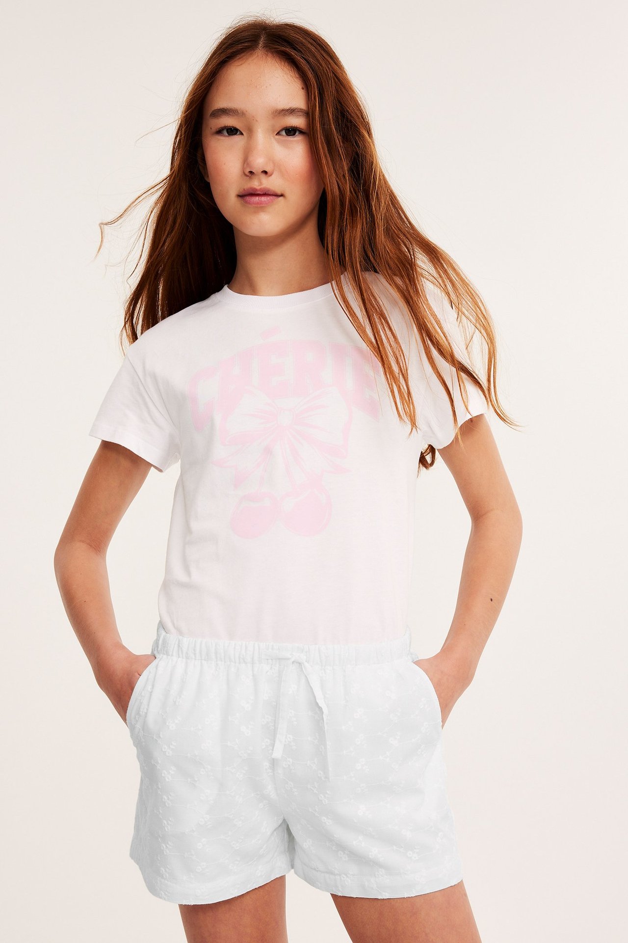 Frontvy: Barn i vita broderade bomullsshorts, dragsko i midjan, och vit t-shirt med rosa tryck.