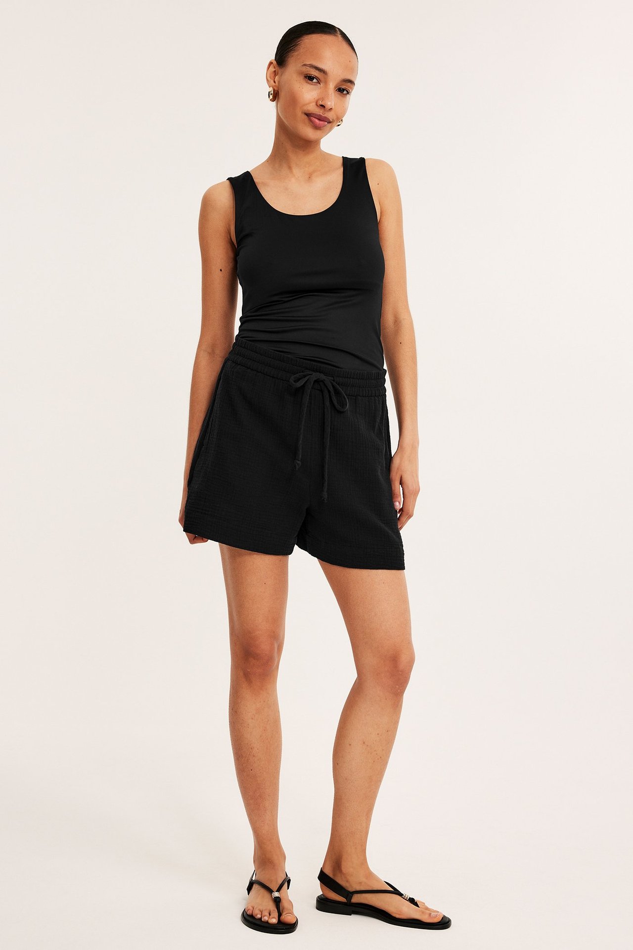 Frontvisning: Kvinne i svarte strukturerte bomullsshorts med knytebånd, svart singlet og svarte sandaler.