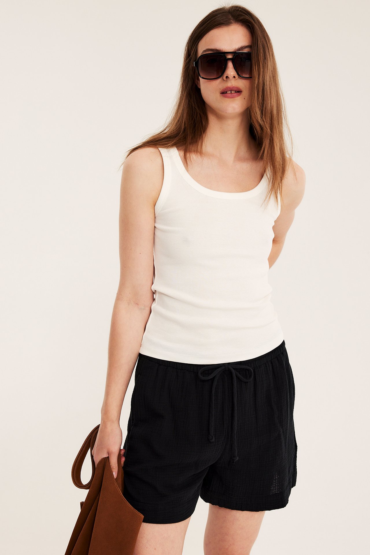 Nainen yllään mustat puuvillashortsit kiristysnauhalla, luonnonvalkoinen toppi, mustat aurinkolasit, pitelee ruskeaa laukkua.