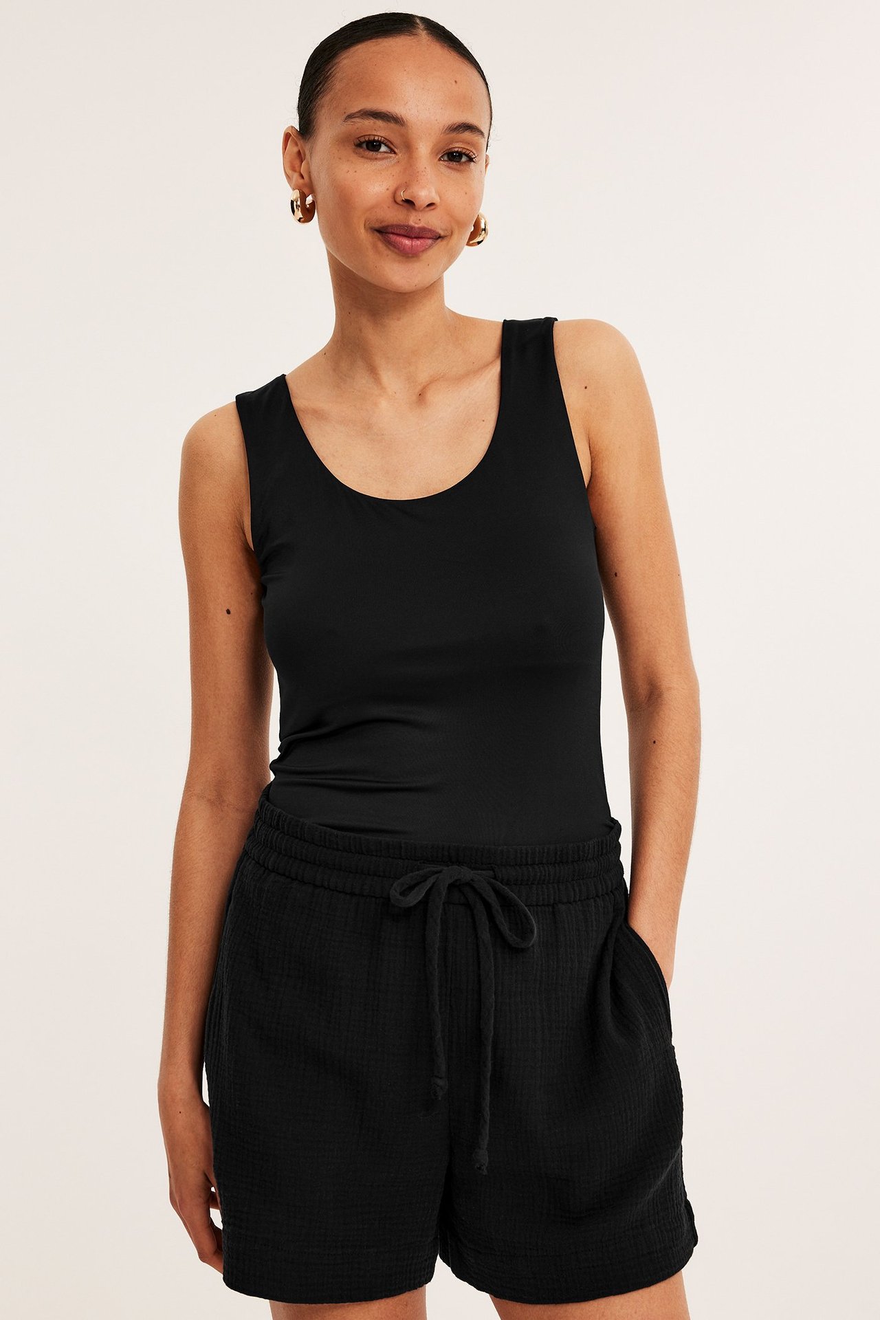 Forfra: kvinne i svarte bomullsgas-shorts med snøring, sidelommer og svart singlet.