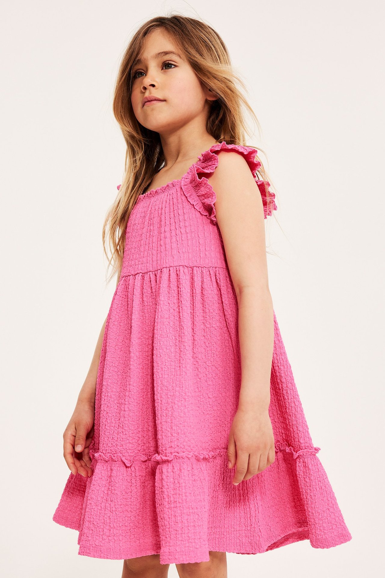 Frontvisning av et barn i en rosa krøllet jerseykjole med volangstropper og etasjeskjørt.