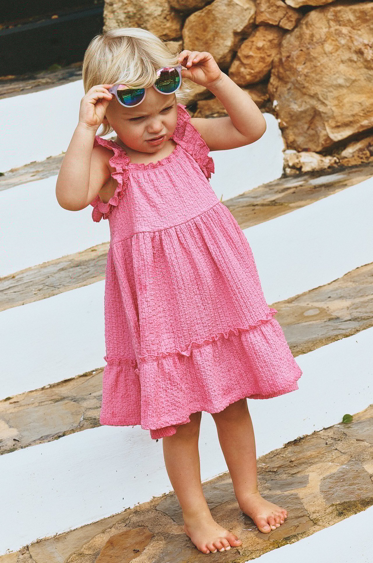 Widok z przodu: dziecko w różowej marszczonej sukience z falbankami, trzyma odblaskowe okulary.