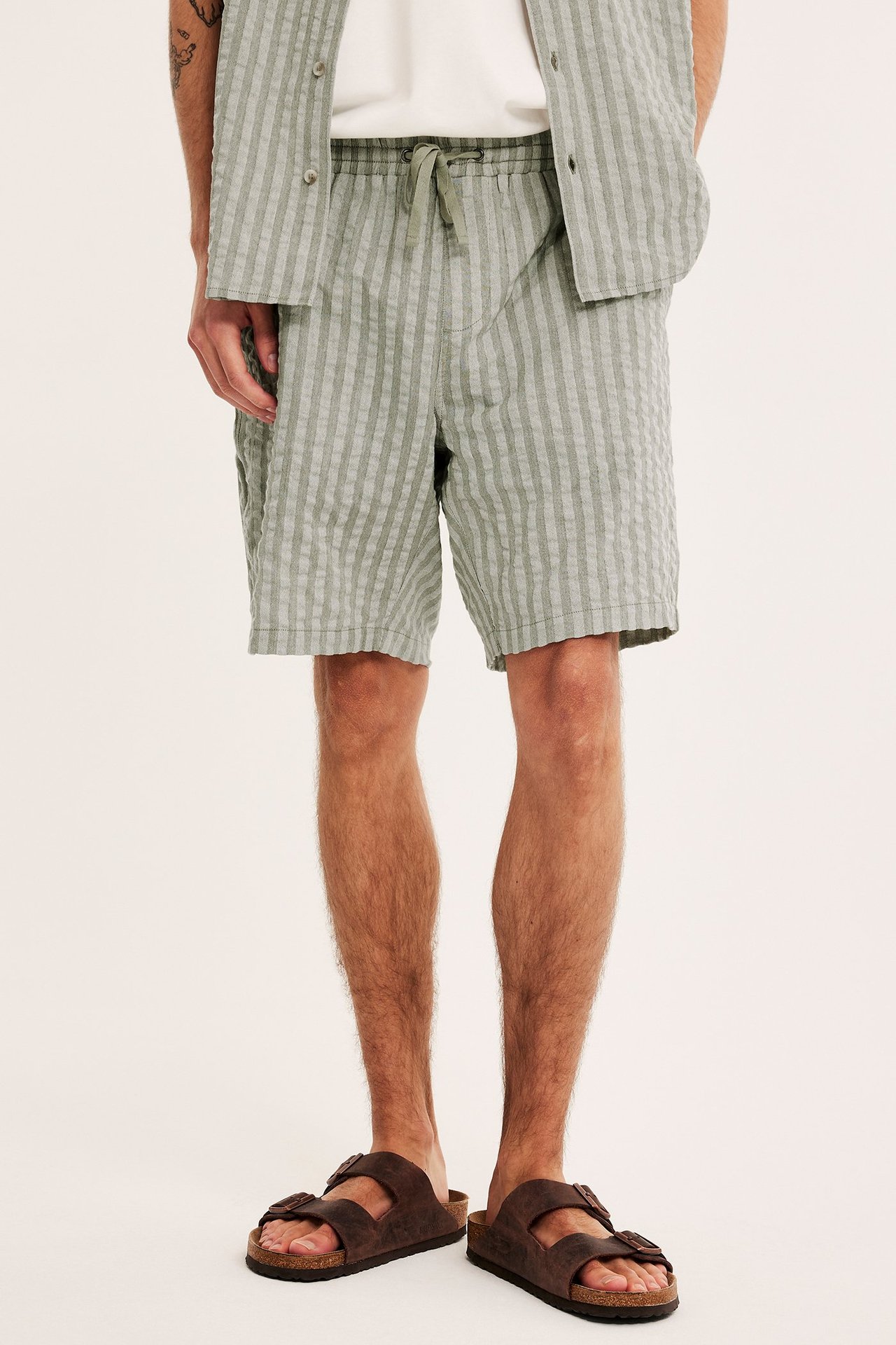 Mies edestä: vihreät raidalliset seersucker-shortsit, yhteensopiva paita, valkoinen t-paita ja ruskeat sandaalit.
