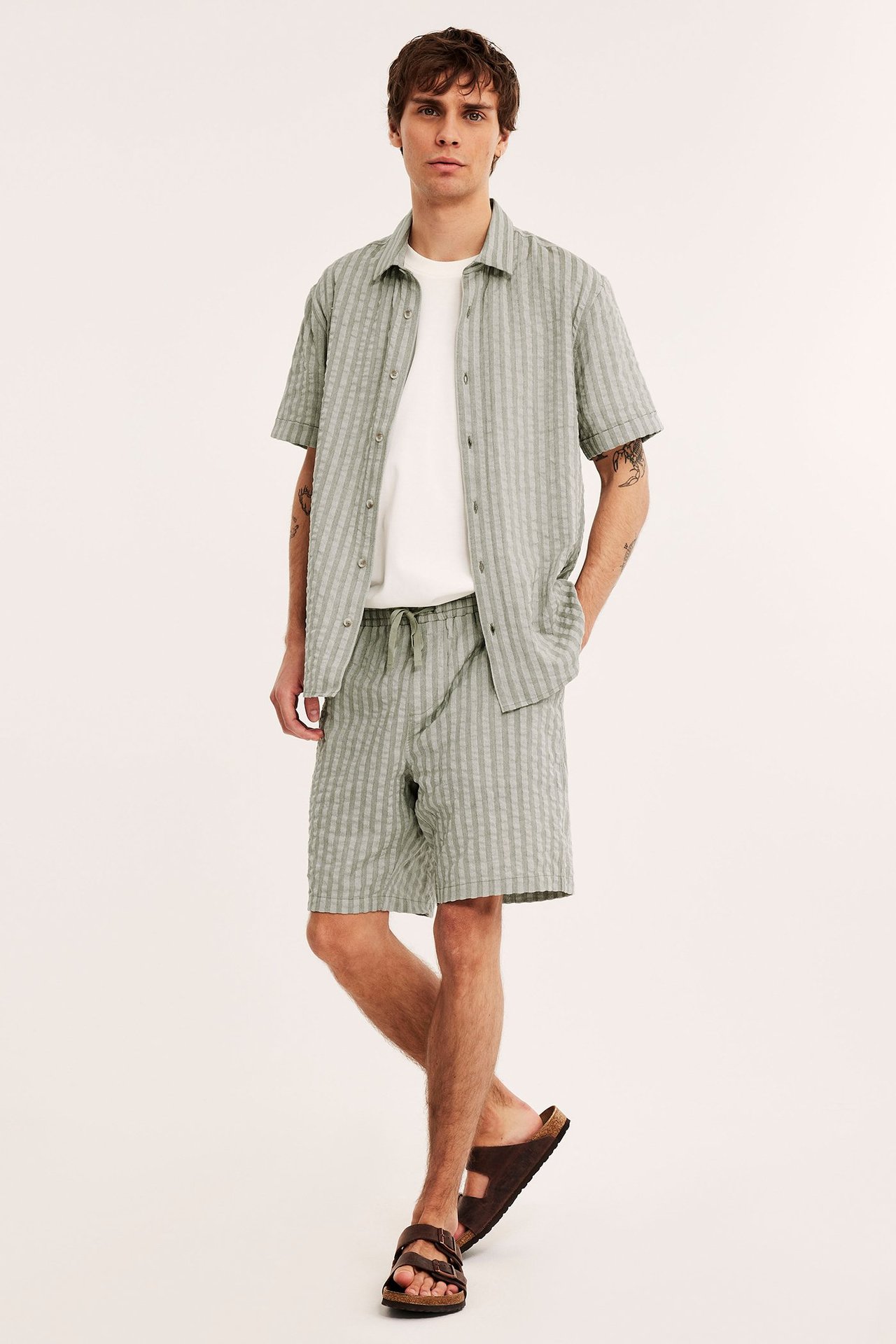 Edestä: mies vihreissä raidallisissa seersucker-shortseissa, yhteensopivassa paidassa, valkoisessa t-paidassa ja ruskeissa sandaaleissa.