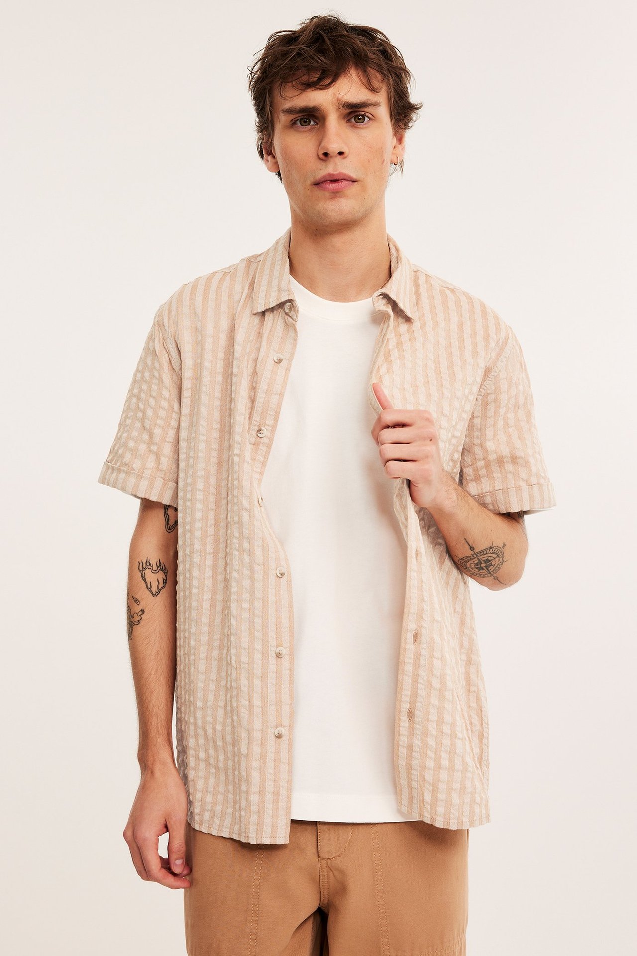 Edestä: Mies avonaisessa beigessä raidallisessa seersucker-lyhythihaisessa paidassa valkoisen t-paidan ja ruskeiden shortsien päällä.