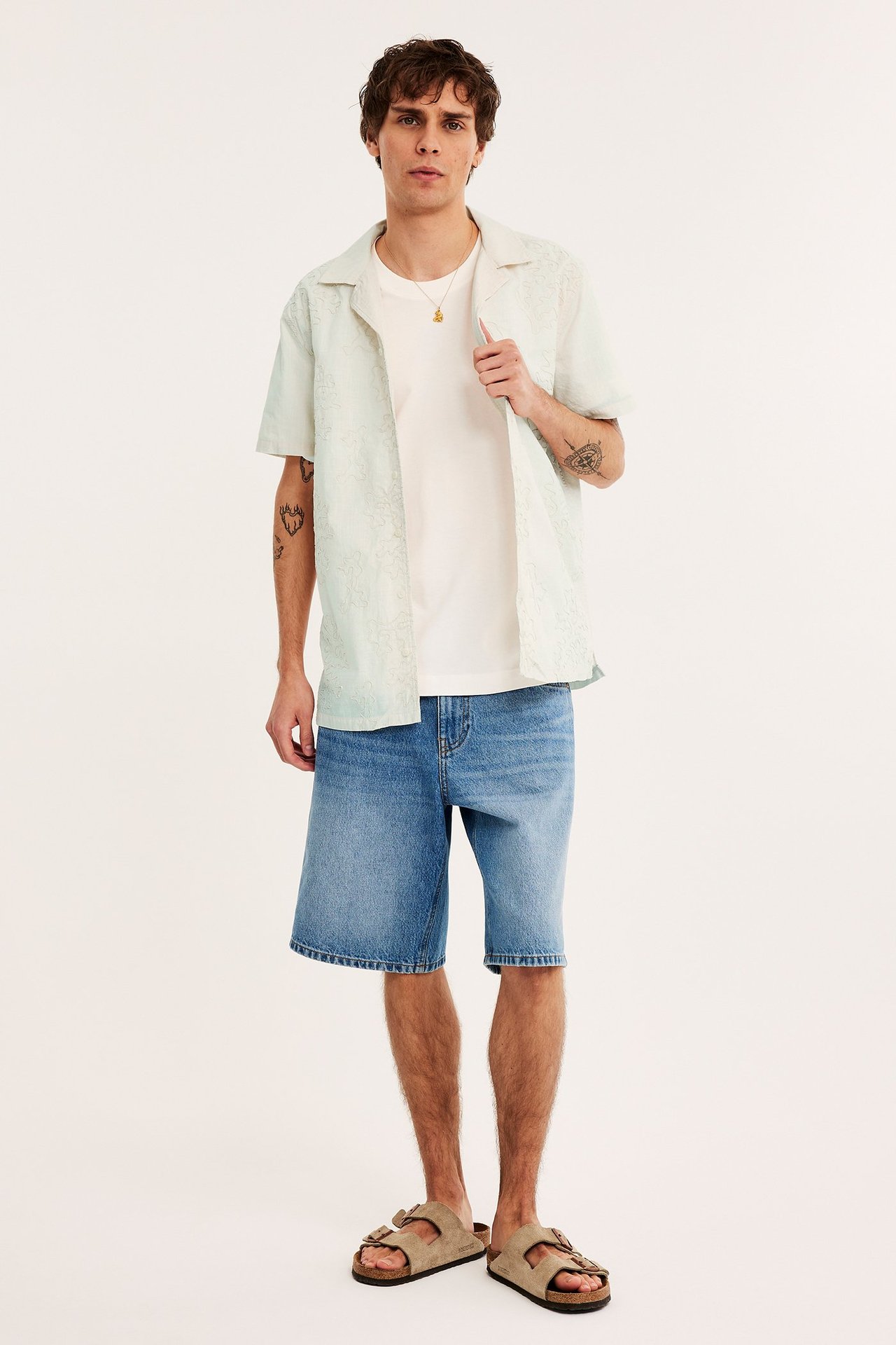 Edestä: mies avoimessa vaaleanbeigessä brodeeratussa puuvillaresorttipaidassa, valkoisessa t-paidassa, sinisissä farkkushortseissa, beigessä sandaaleissa.