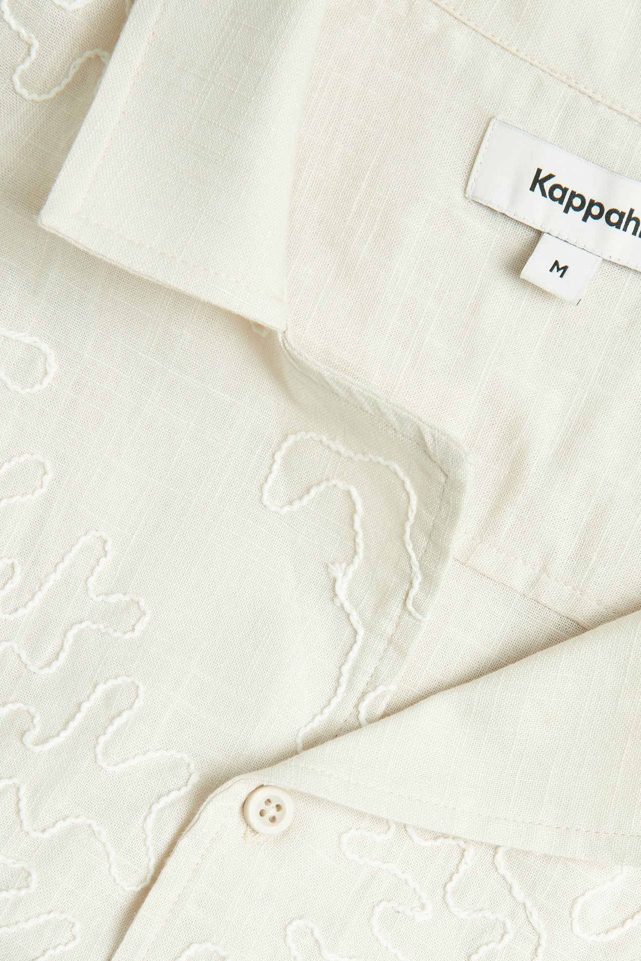 Zbliżenie jasnobeżowej bawełnianej koszuli resortowej z ton w ton haftowanymi falistymi wzorami i guzikiem.