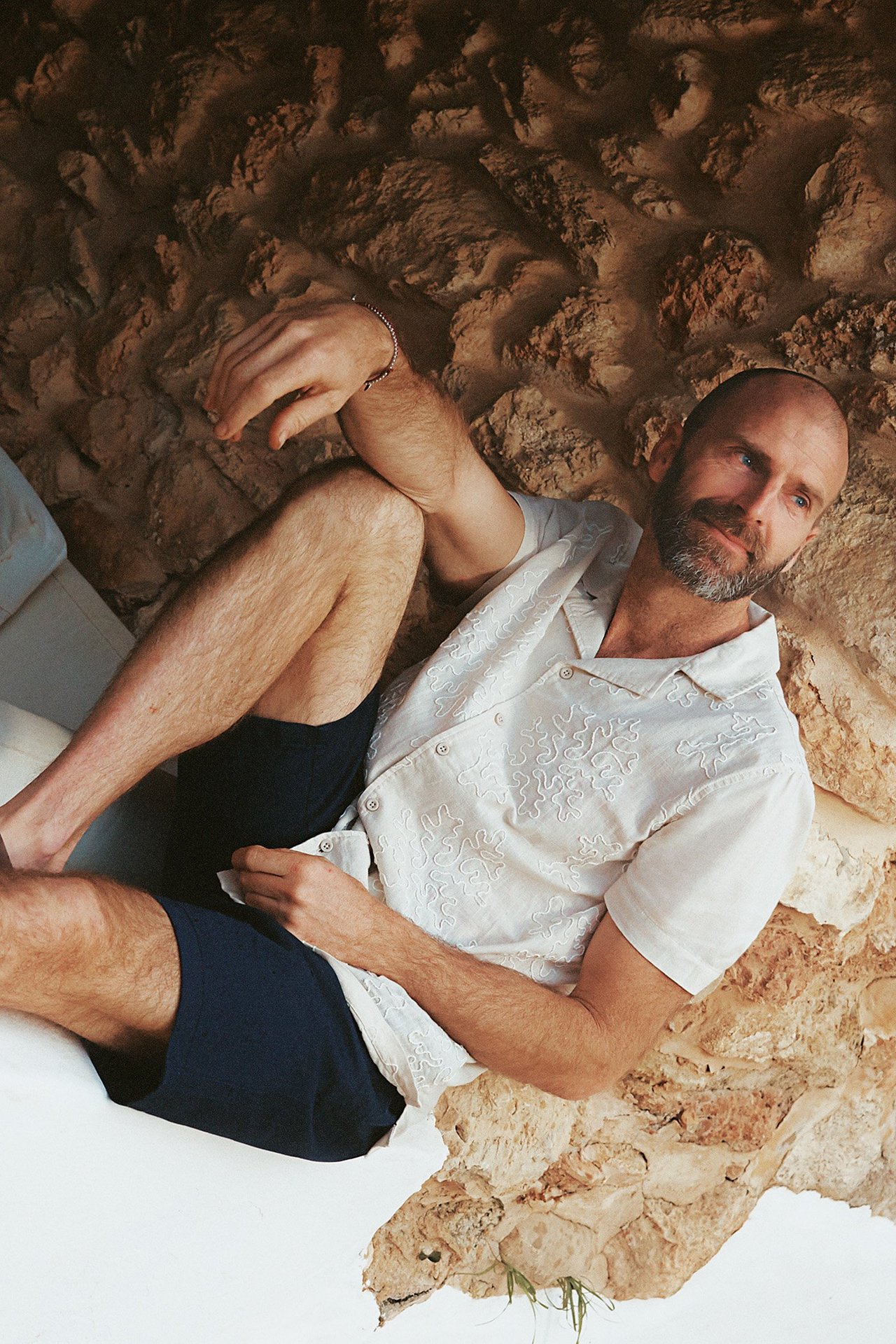 Edestä: Mies vaaleanbeigessä kirjaillussa lyhythihaisessa puuvillapaidassa ja tummansinisissä shortseissa.