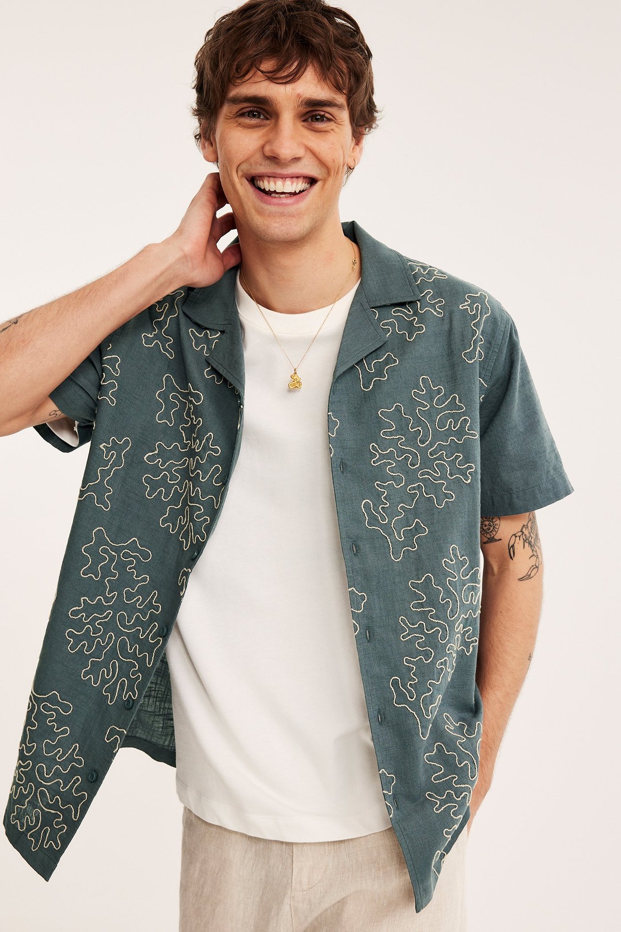 Mies tummanharmaassa puuvillaisessa lyhythihaisessa paidassa valkoisella kirjonnalla, valkoisen t-paidan päällä.