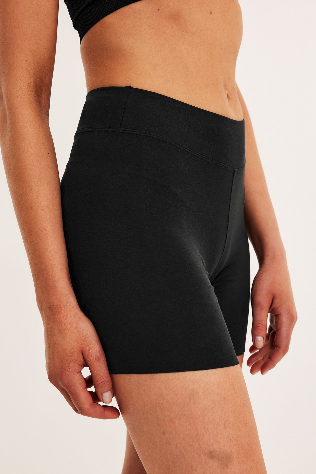 Sidebilde av en kvinne i svarte høytlivs shorts-truser i bomull og en svart BH-topp.