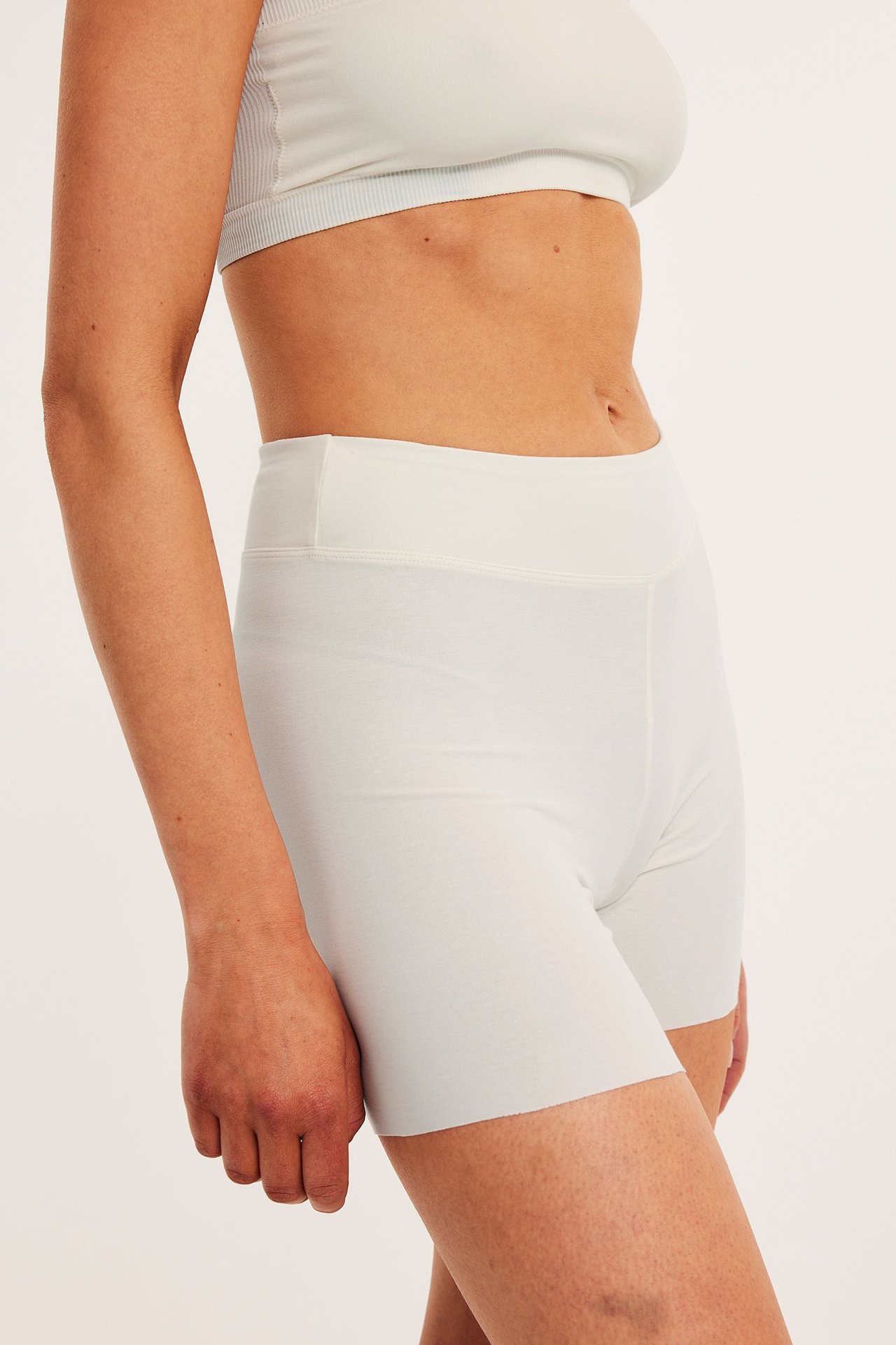 Sidebilde av kvinne i hvite, høytlivs bomullsshortstruser og en lys grå ribbet bralette.