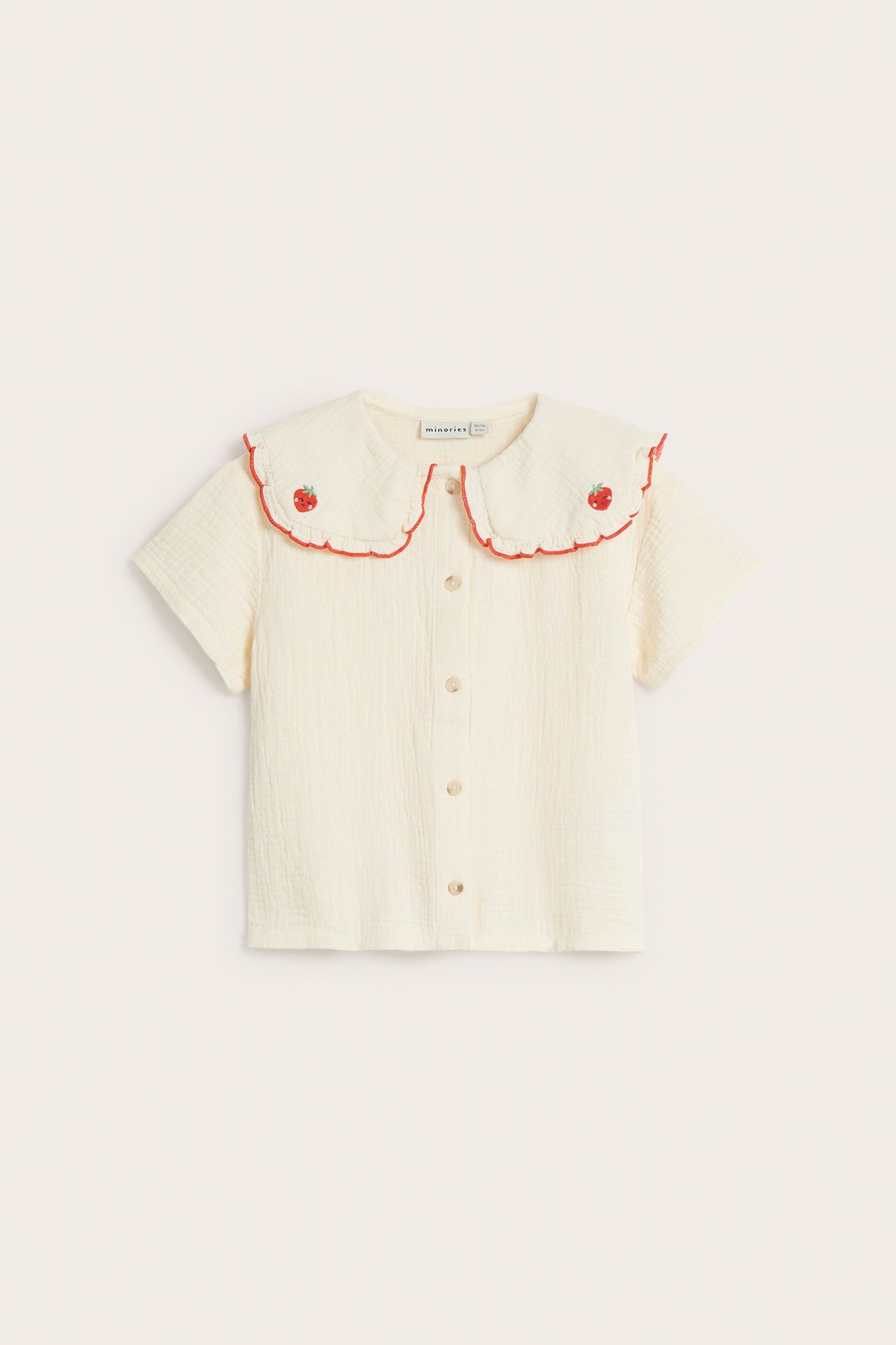 Forfra: Barnebluse i beige musselin med korte ermer, knapper foran og brodert jordbærkrage.