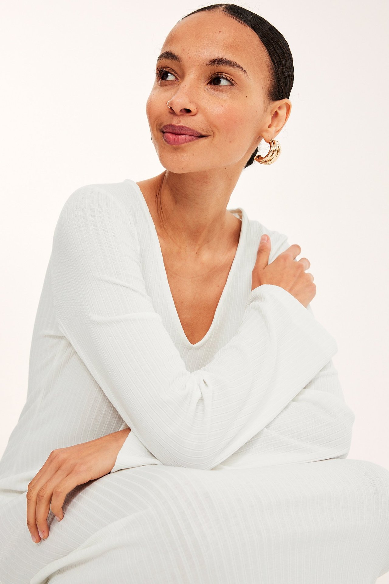 Nærbilde av kvinne i offwhite ribbet V-hals langermet kjole og gullfargede ringøredobber.