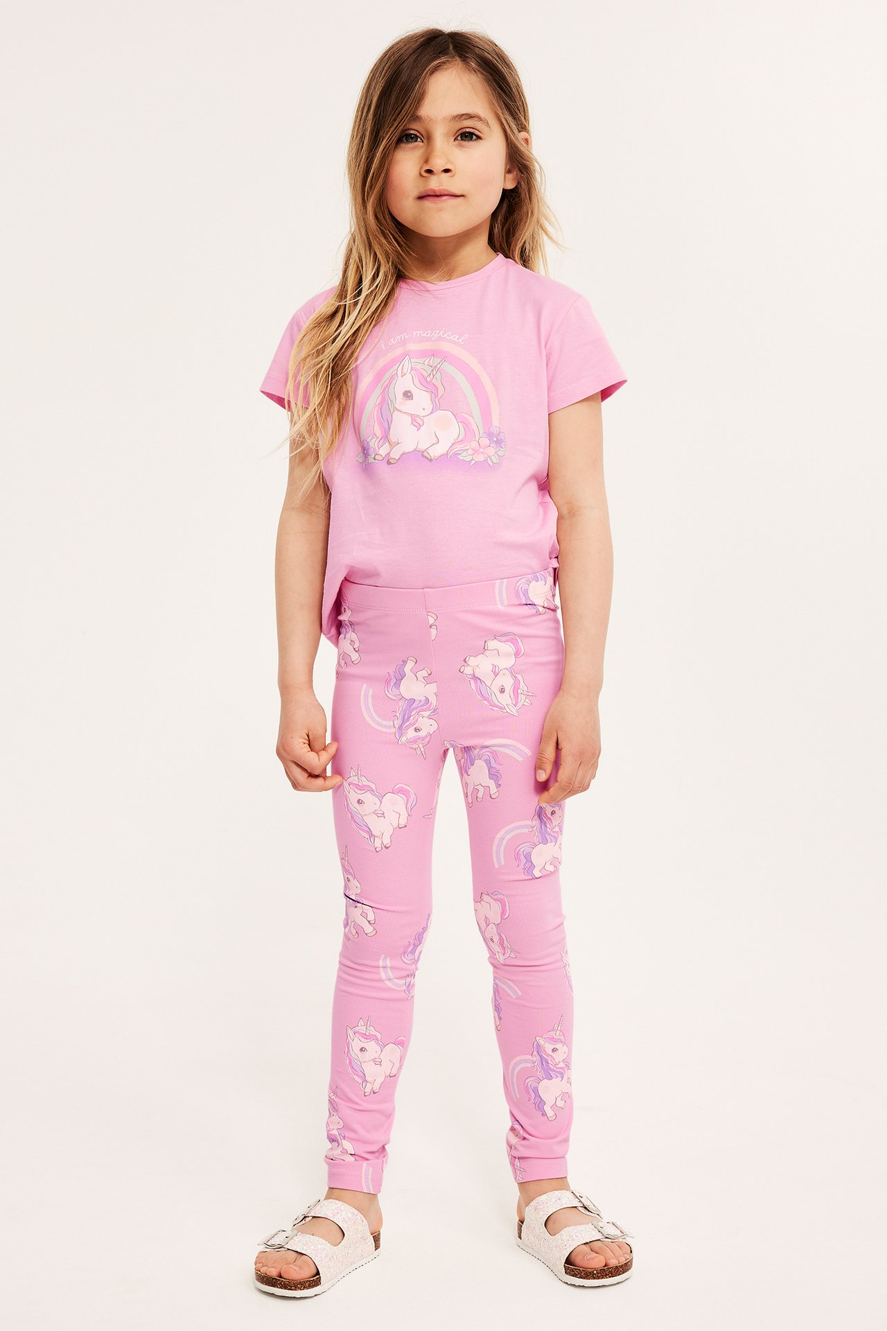 Framifrån: ett barn i rosa leggings med enhörningsmönster, en rosa enhörningströja och vita sandaler.
