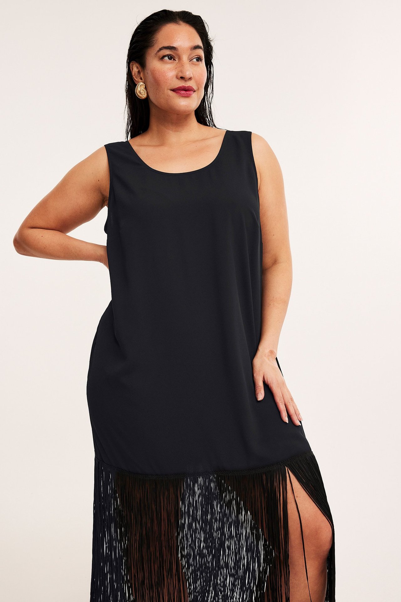 Edestäpäin: nainen mustassa hihattomassa polyesterimekossa, jossa pitkä hapsuhelma ja kultaiset spiraalikorvakorut.