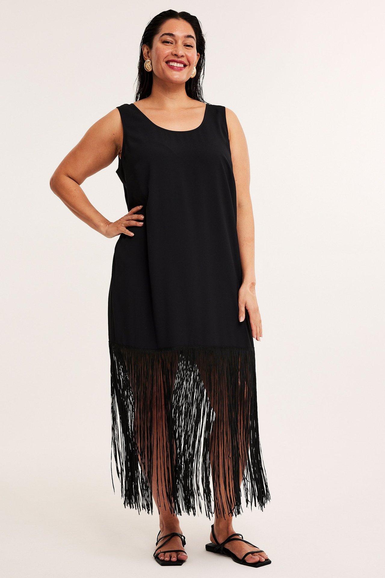 Edestä kuvattu nainen mustassa hihattomassa polyesterimekossa, jossa on pitkä hapsuhelma ja mustat sandaalit.