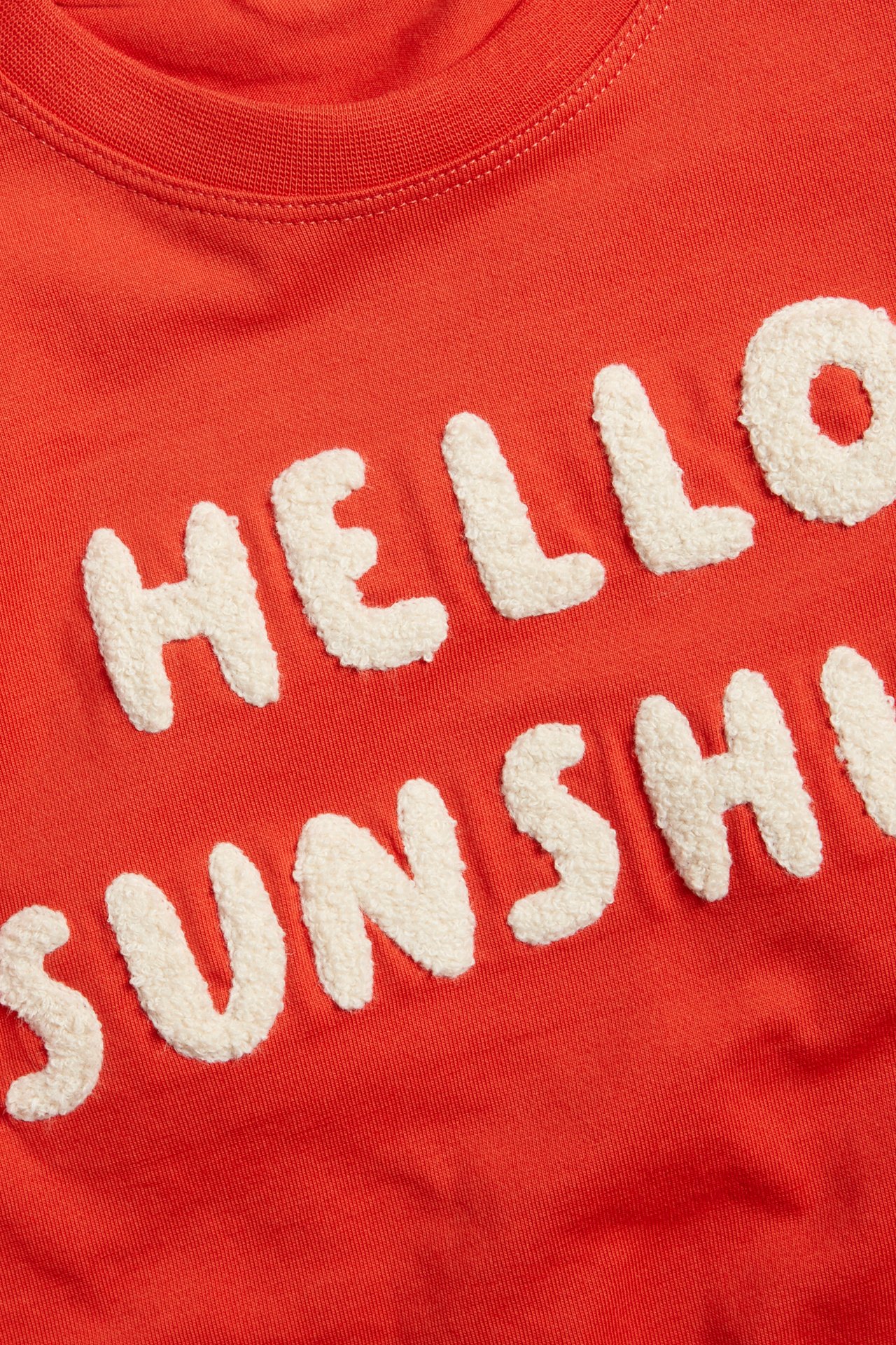 Närbild på en röd bomulls-t-shirt med vit frottéapplikation "HELLO SUNSHINE" framtill.