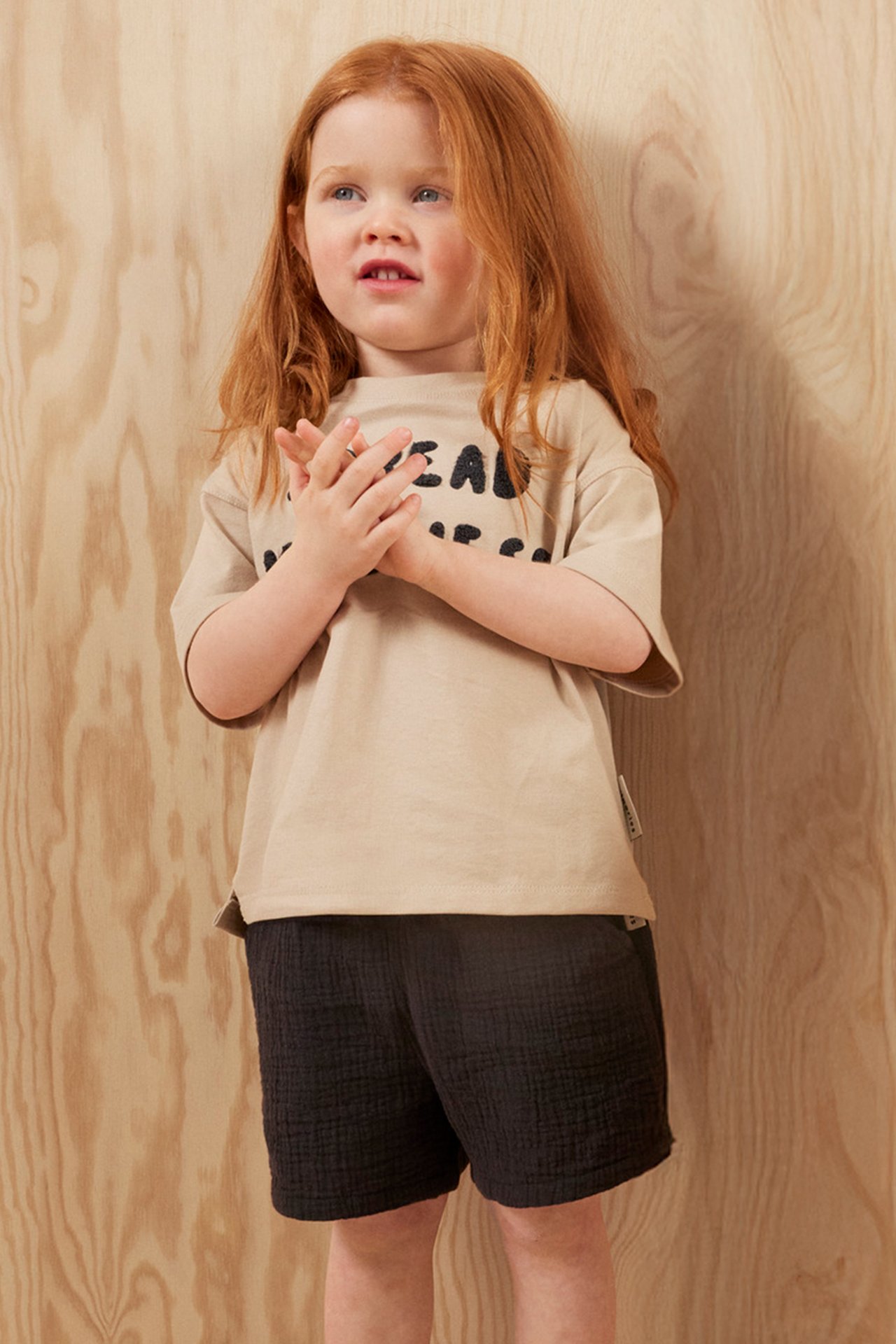Framifrån: ett barn i ljusbeige bomulls-t-shirt med svart frottéapplikation och mörka texturerade shorts.
