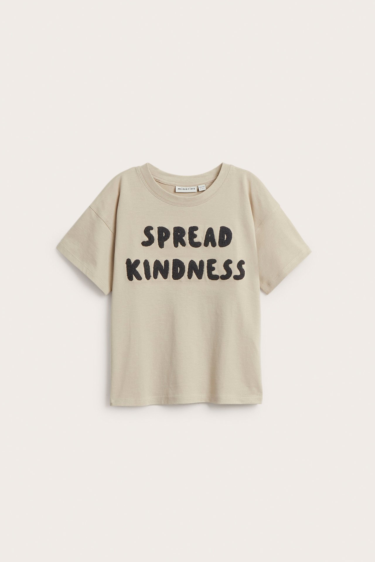 Frontvy av en ljusbeige bomulls-t-shirt med svart 'SPREAD KINDNESS' frottéapplikation.