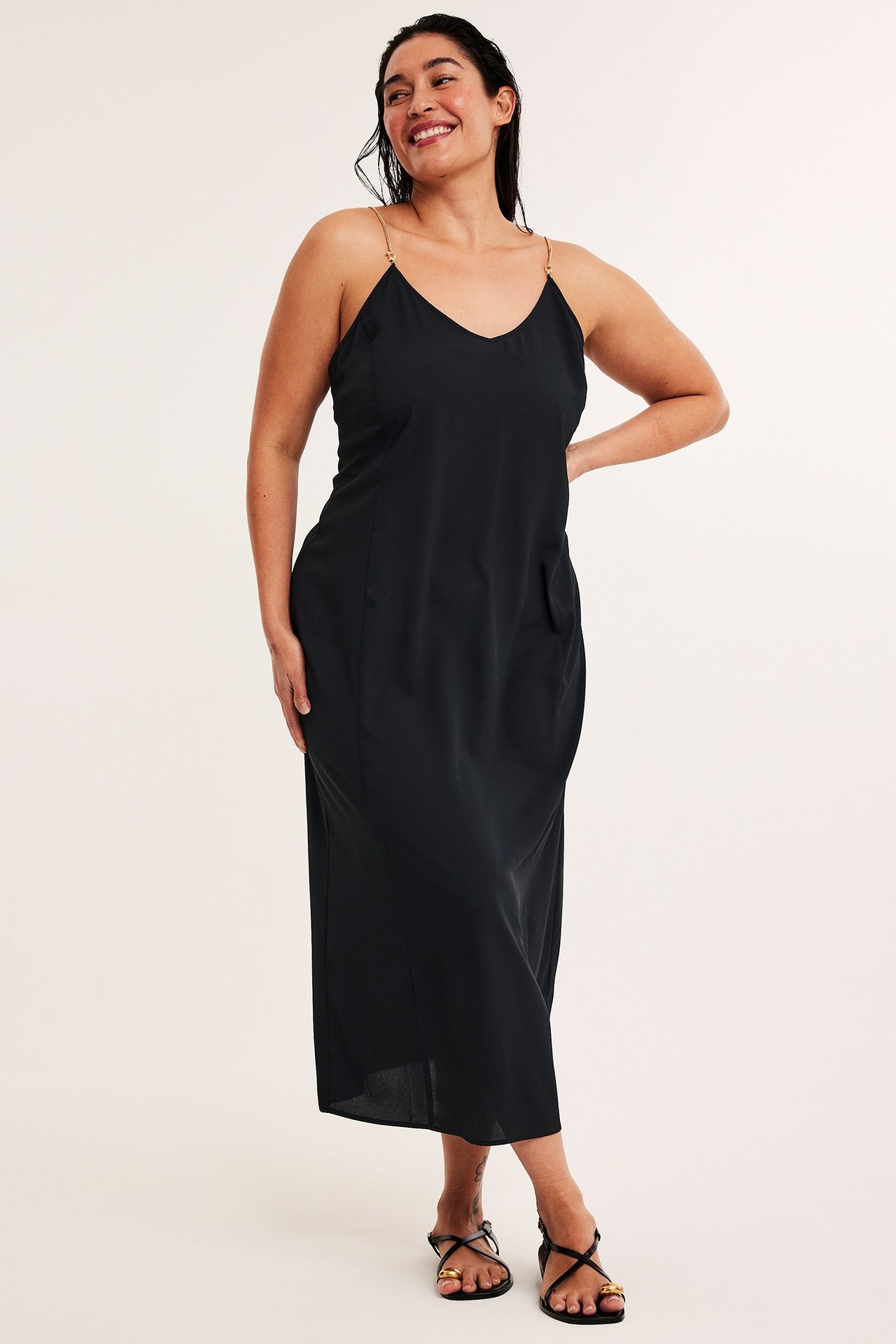 Edestäpäin: nainen mustassa midimekossa, jossa kultaiset ketjuolkaimet, ja mustat sandaalit.