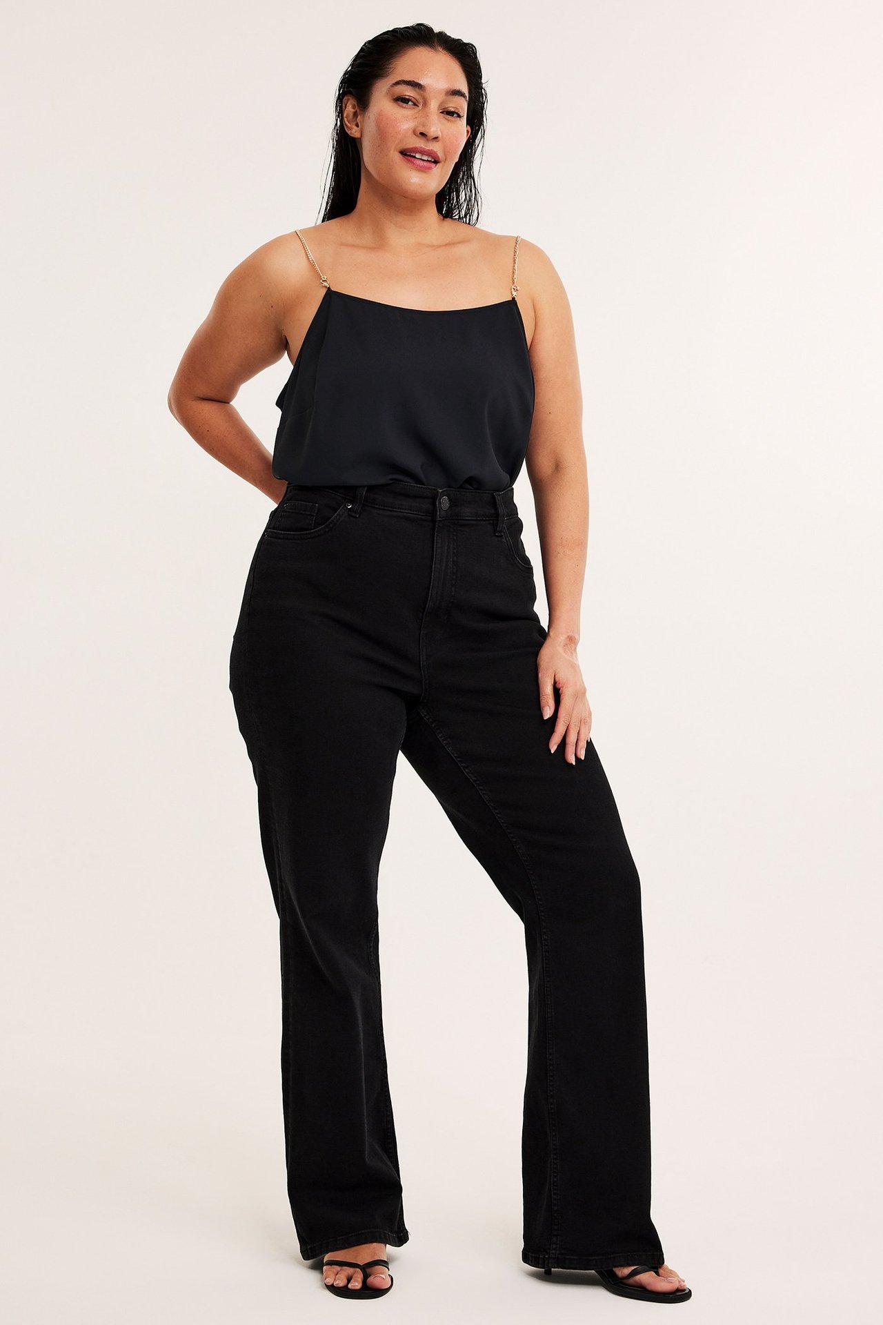 Forfra: kvinne i svart polyestertopp med gullfargede kjedestropper og svarte jeans.