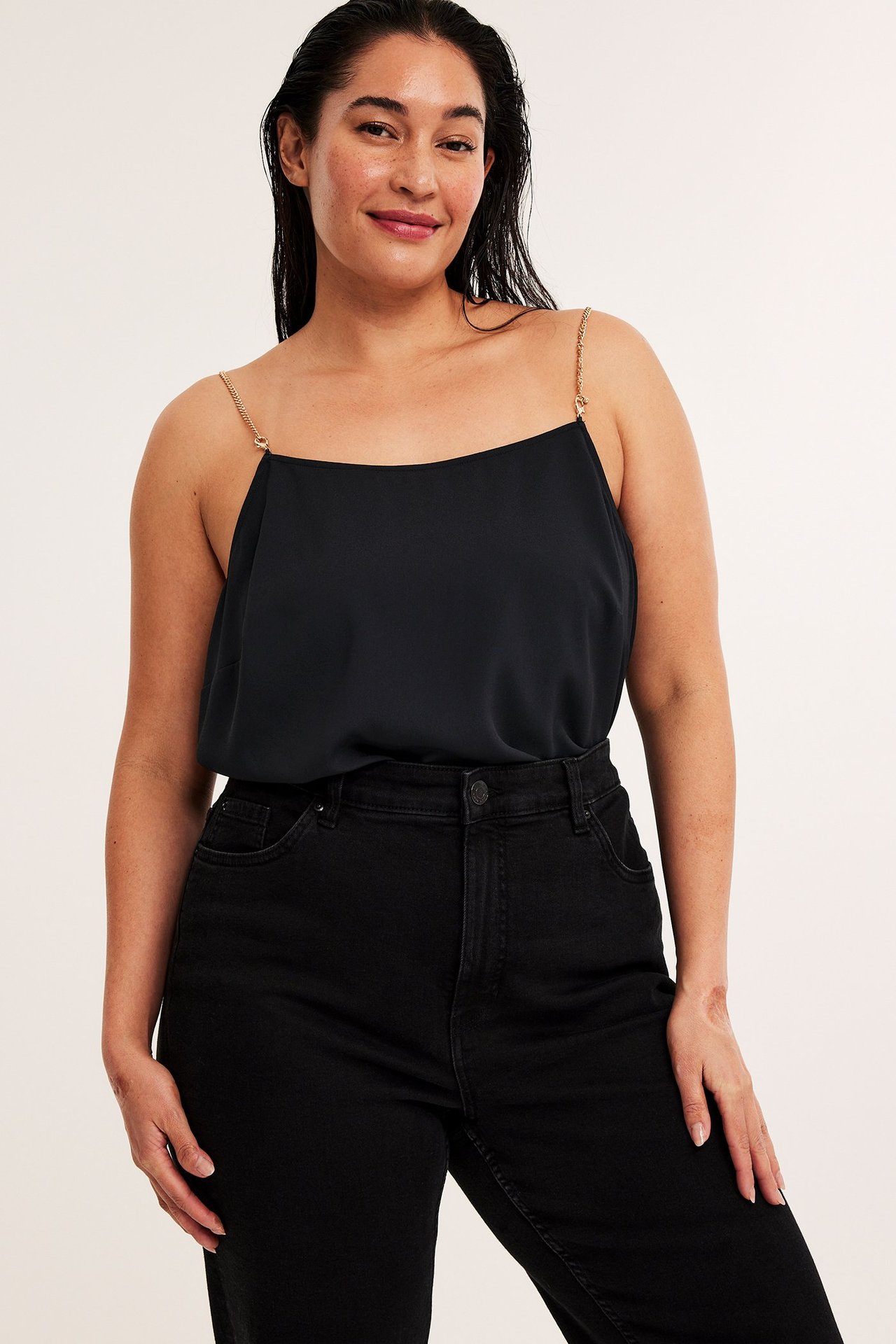 Forfra: kvinne i svart polyestertopp med gullfargede kjedestropper og svarte jeans.