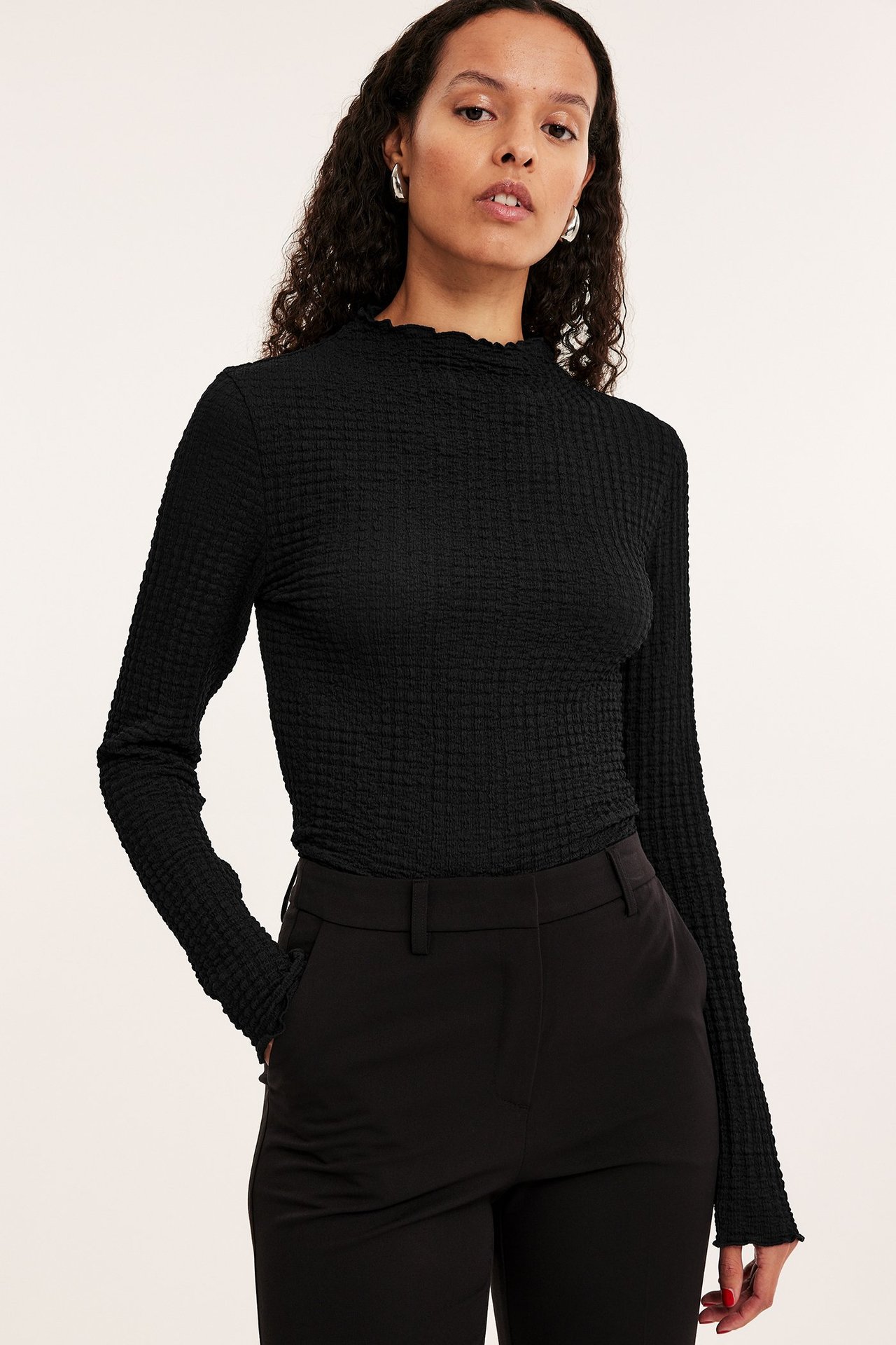 Widok z przodu kobiety w czarnym, strukturalnym topie z długim rękawem, półgolfem i czarnych spodniach.