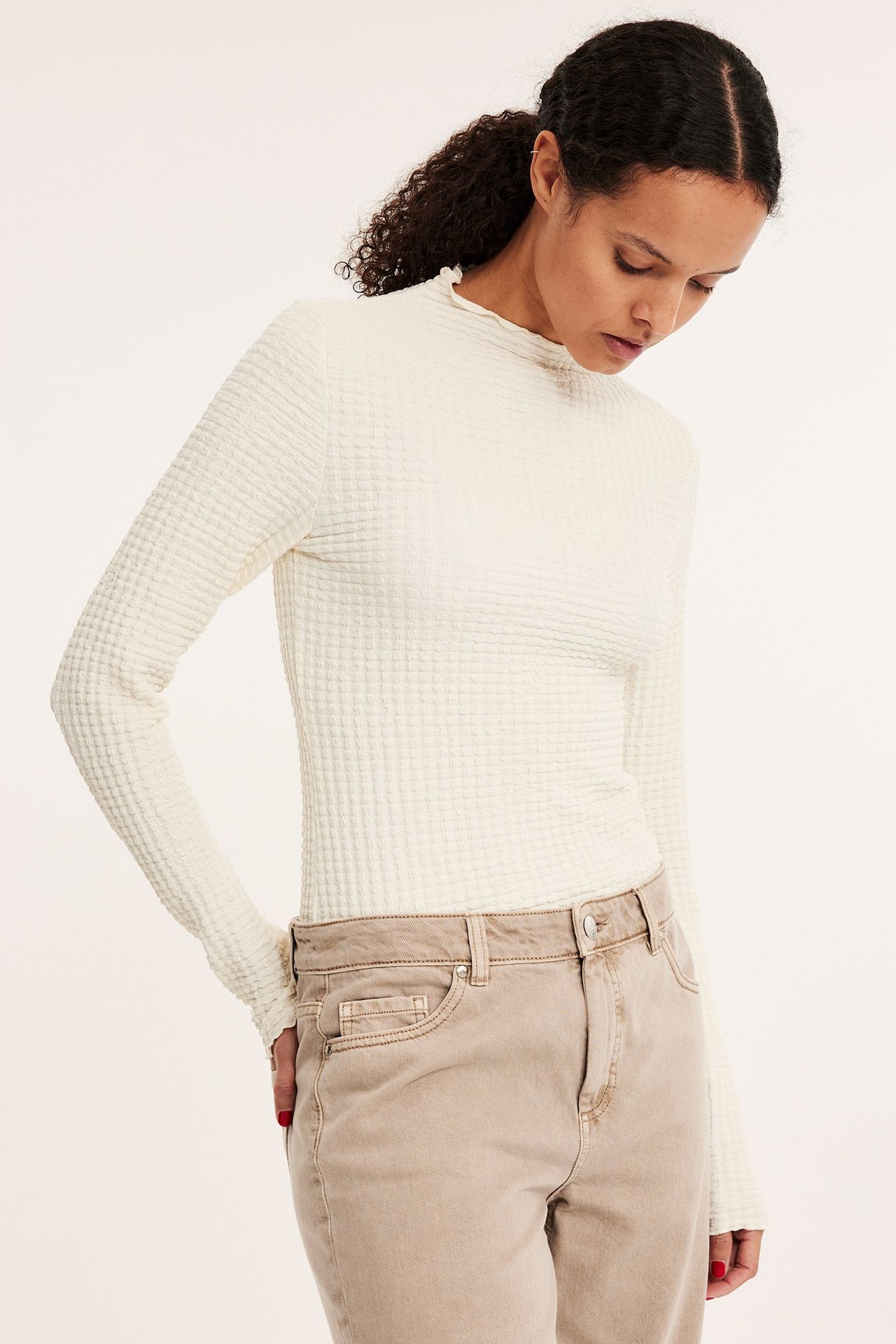 Widok z przodu: Kobieta w kremowym, strukturalnym topie z długim rękawem i półgolfem, w beżowych spodniach.