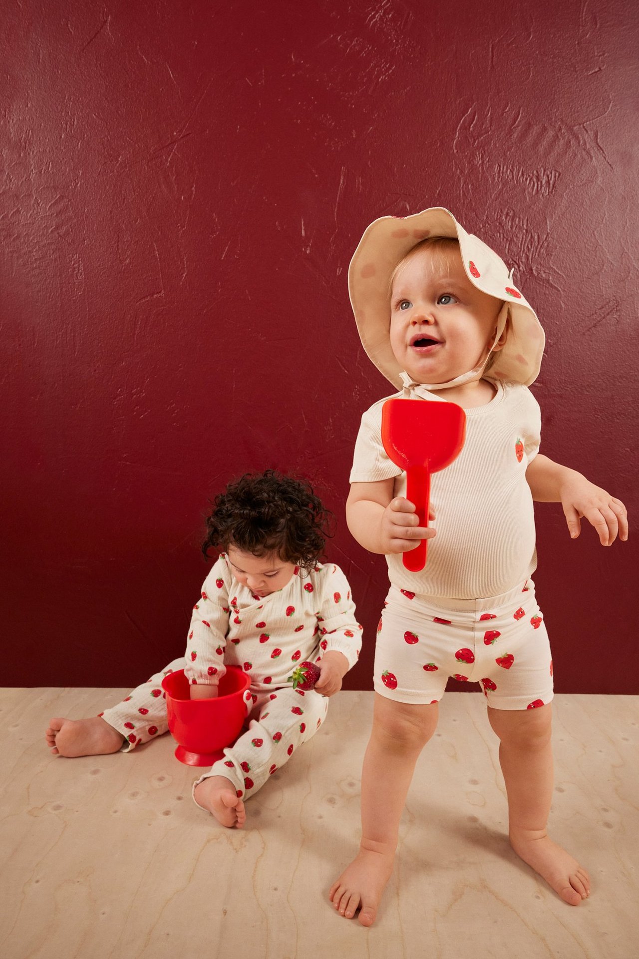 Vauva luonnonvalkoisissa ribatuissa mansikkashortseissa, yhteensopivassa topissa ja hatussa. Toinen vauva samassa kuosissa.