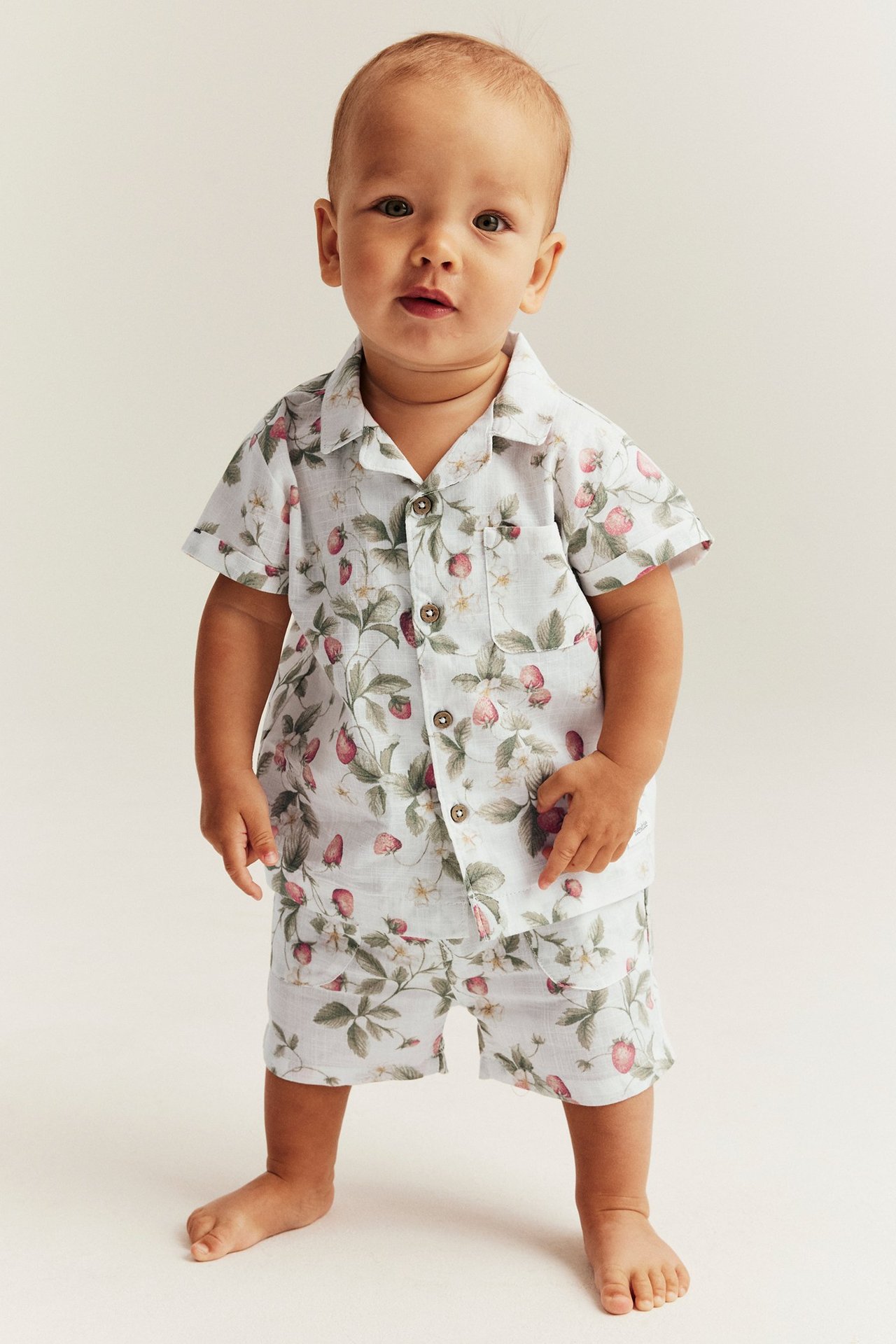 Edestä: vauva yllään luonnonvalkoiset puuvillashortsit ja yhteensopiva mansikkakuvioinen paita.