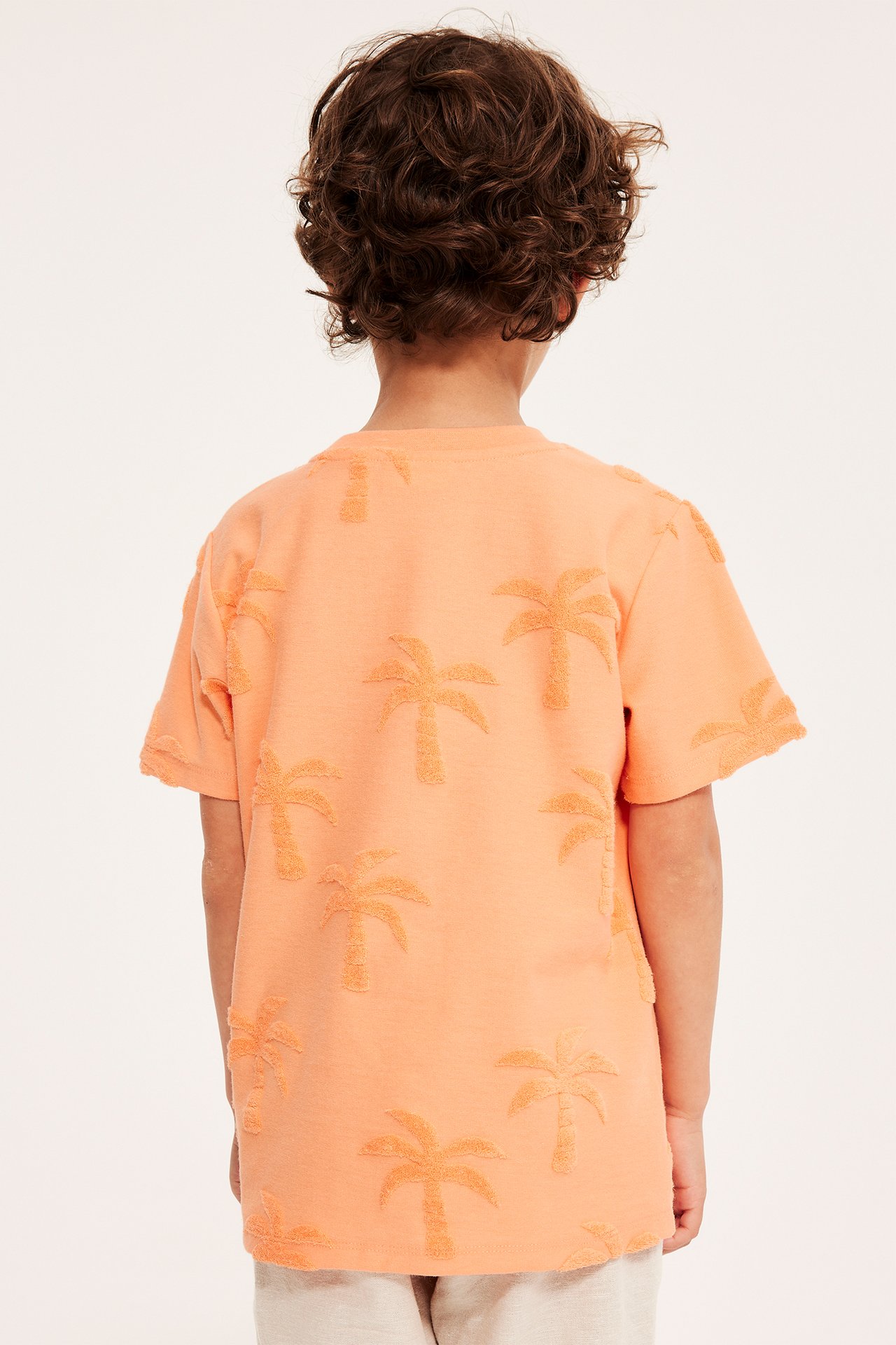 Bakifrån: ett barn i orange t-shirt med frottépalmmönster och ljusa shorts.