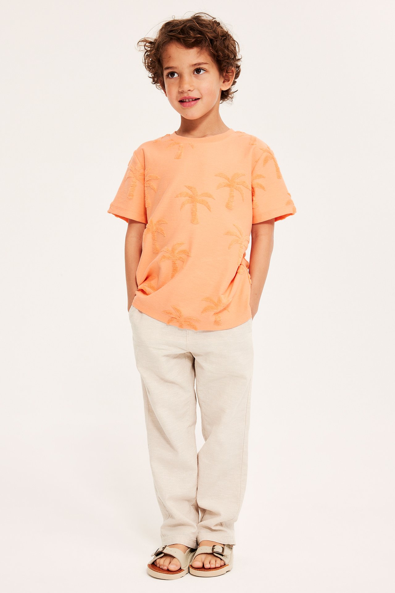 Framifrån, ett barn bärande en orange t-shirt med frottépalmmönster, ljusbeige byxor och beiga sandaler.