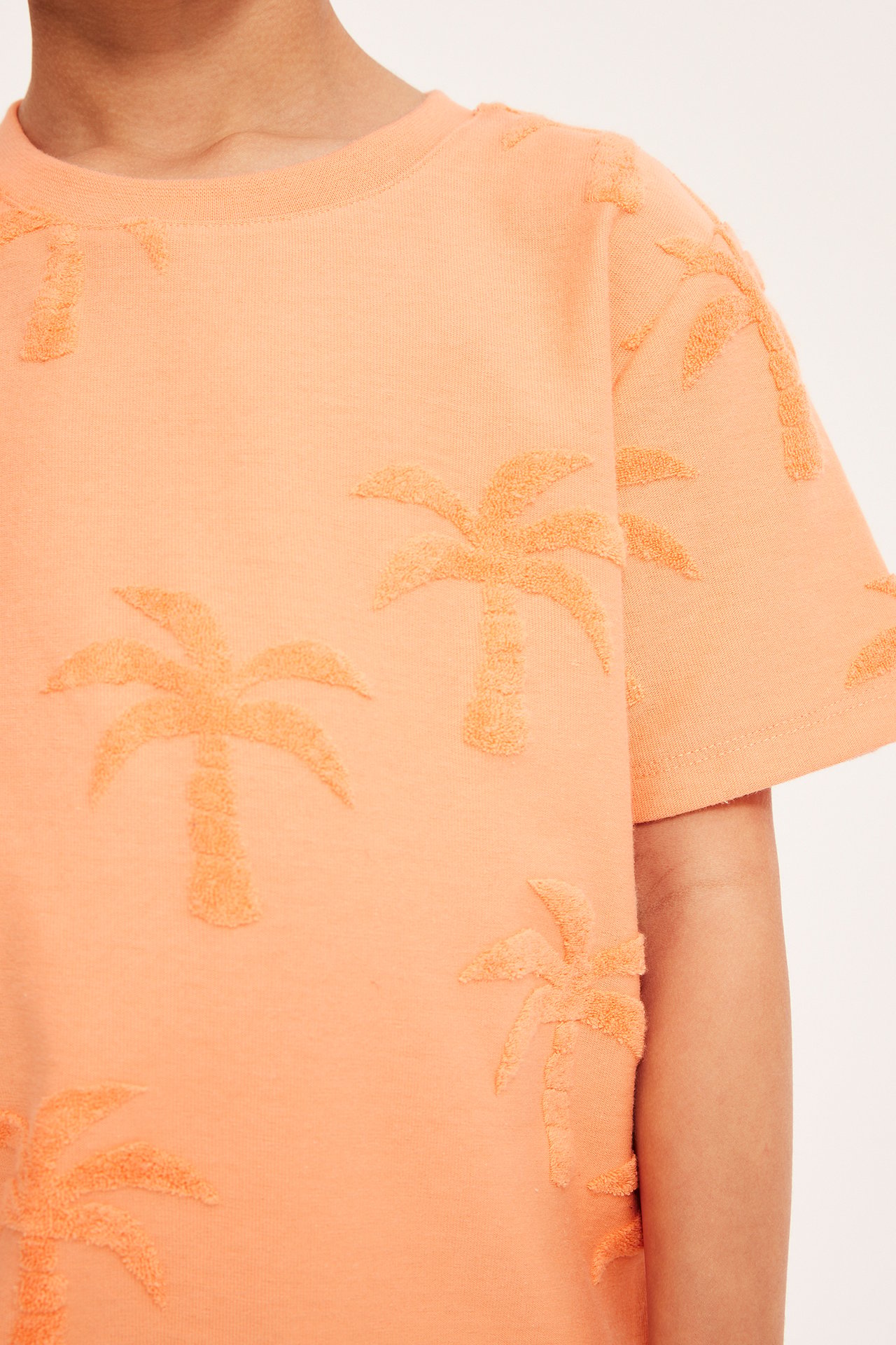 Närbild på ett barn i en orange t-shirt med ribbad rundhals och frottépalm-mönster.