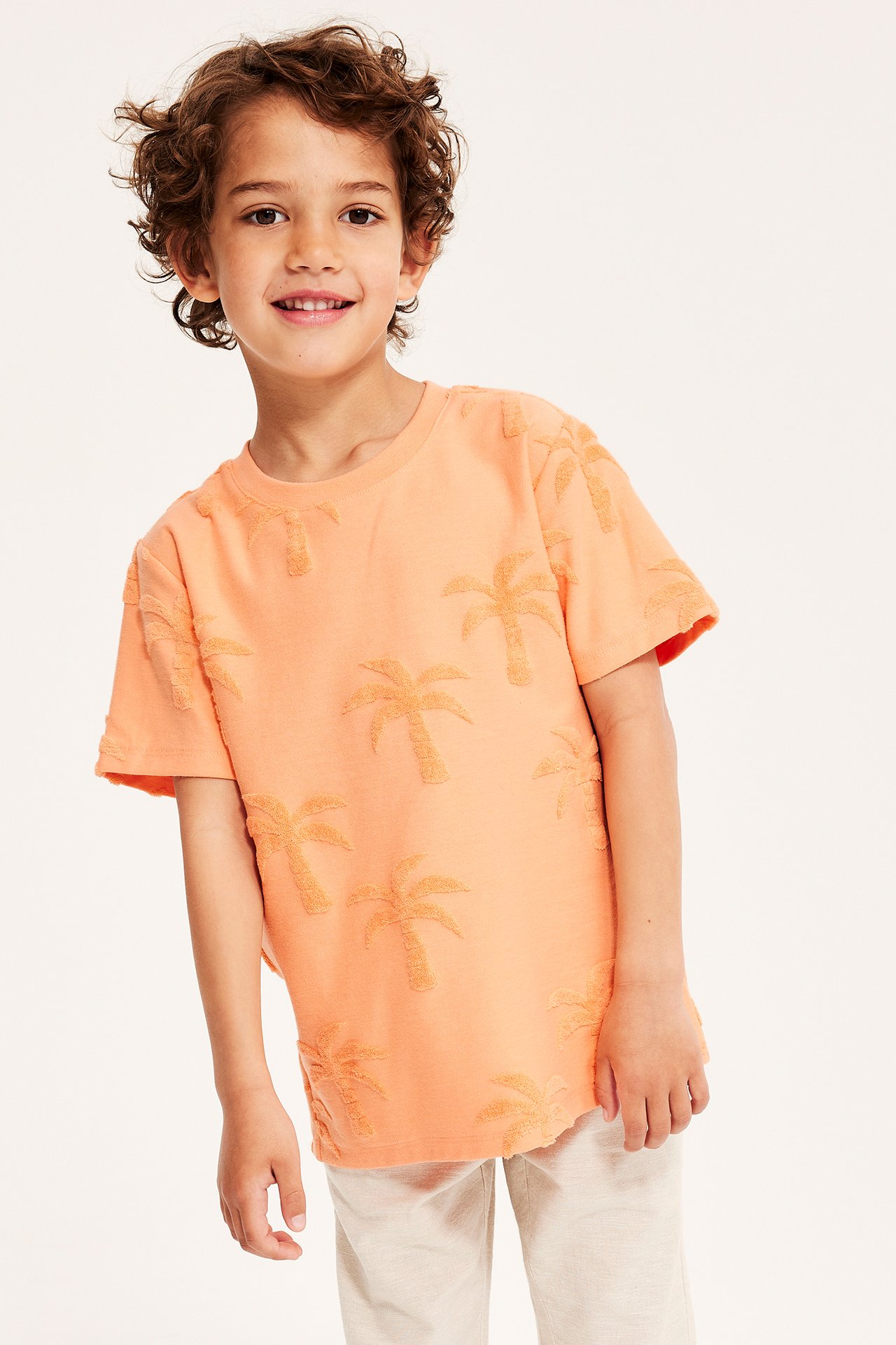 Framifrån: ett barn i orange kortärmad T-shirt med frottépalmmönster och ljusbeige byxor.