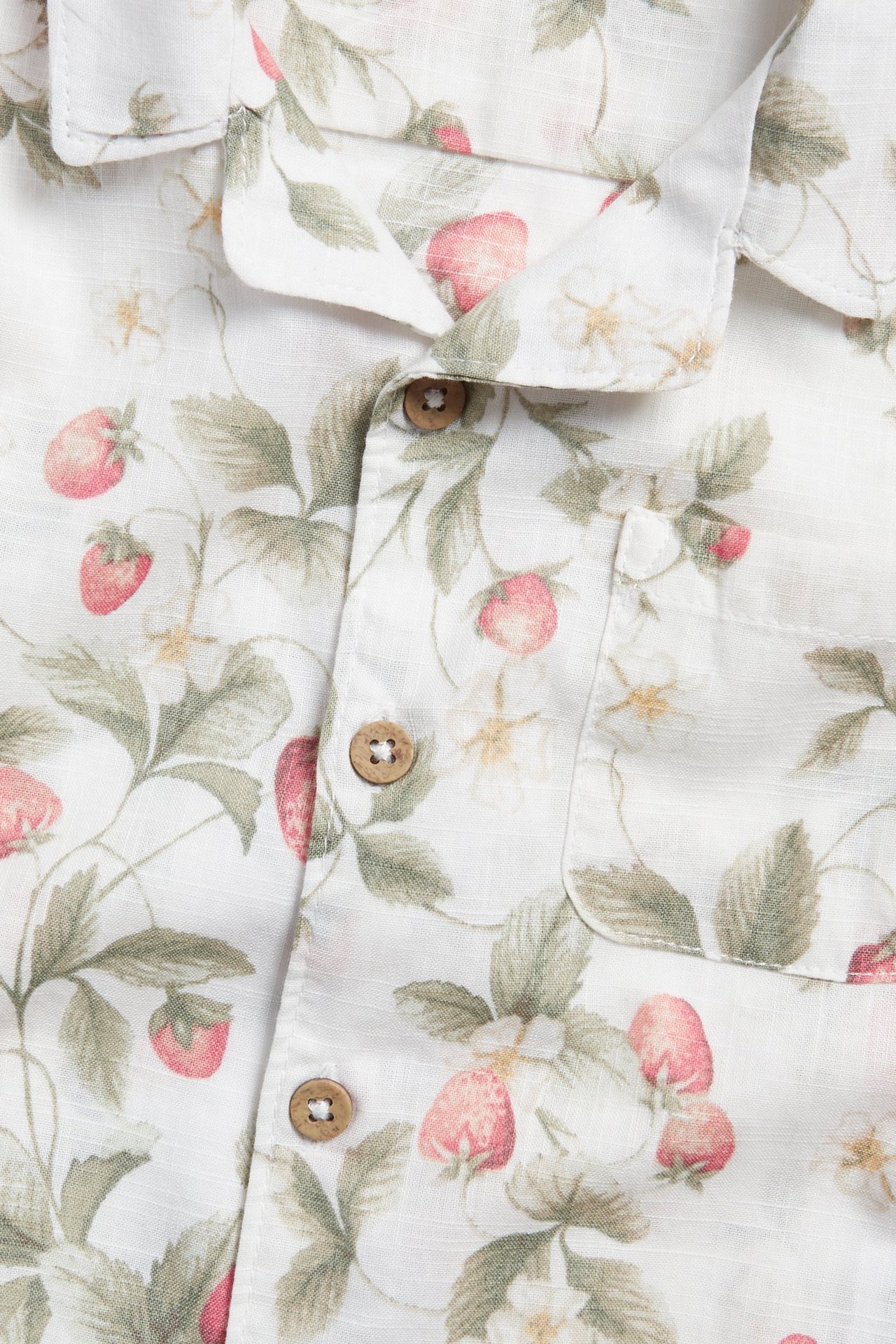 Zbliżenie z przodu: ecru bawełniana koszula z czerwonym wzorem truskawek, kołnierzem i drewnianymi guzikami.