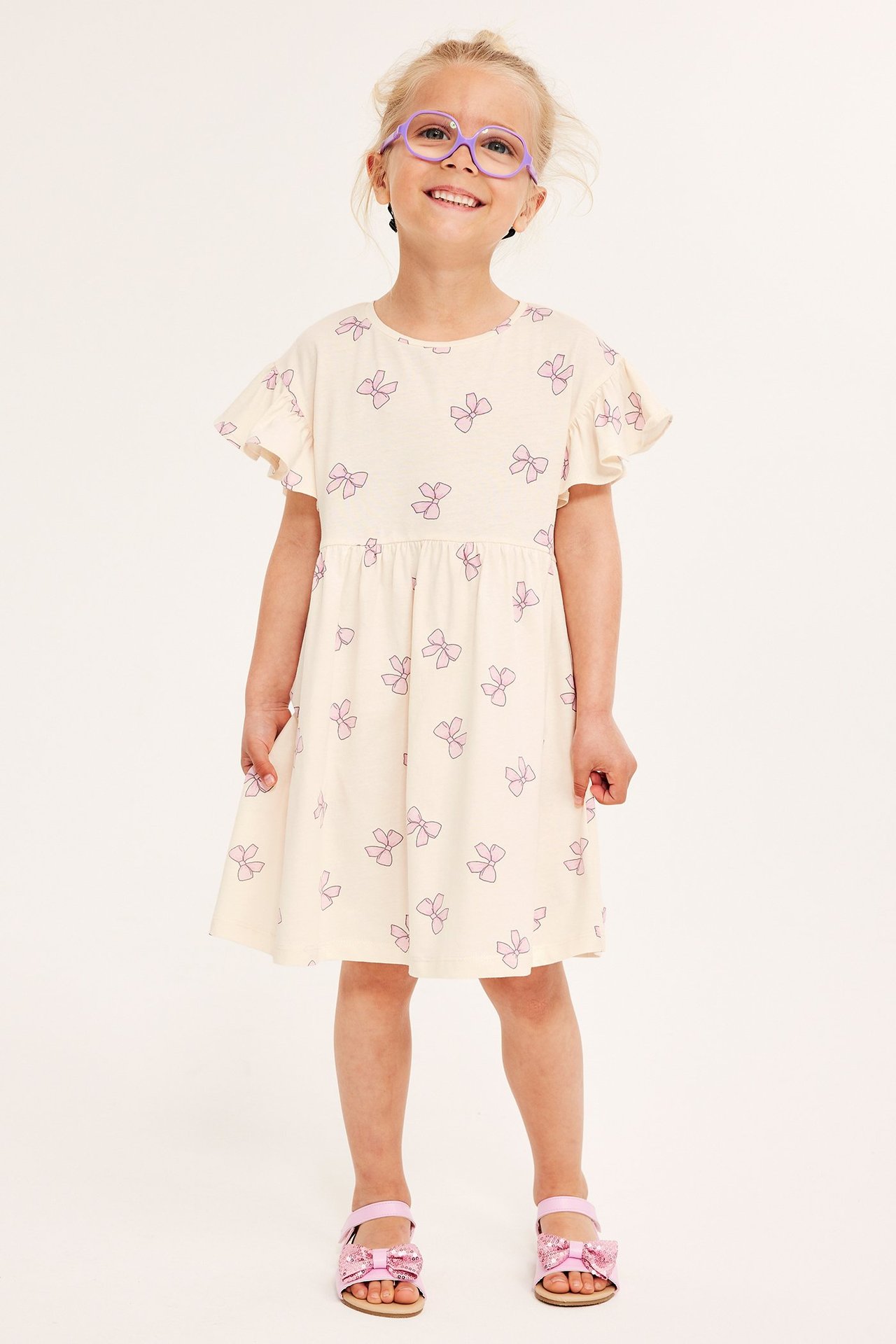 Widok z przodu dziecka w ecru bawełnianej sukience w fioletowe kokardki, fioletowych okularach i różowych sandałach.