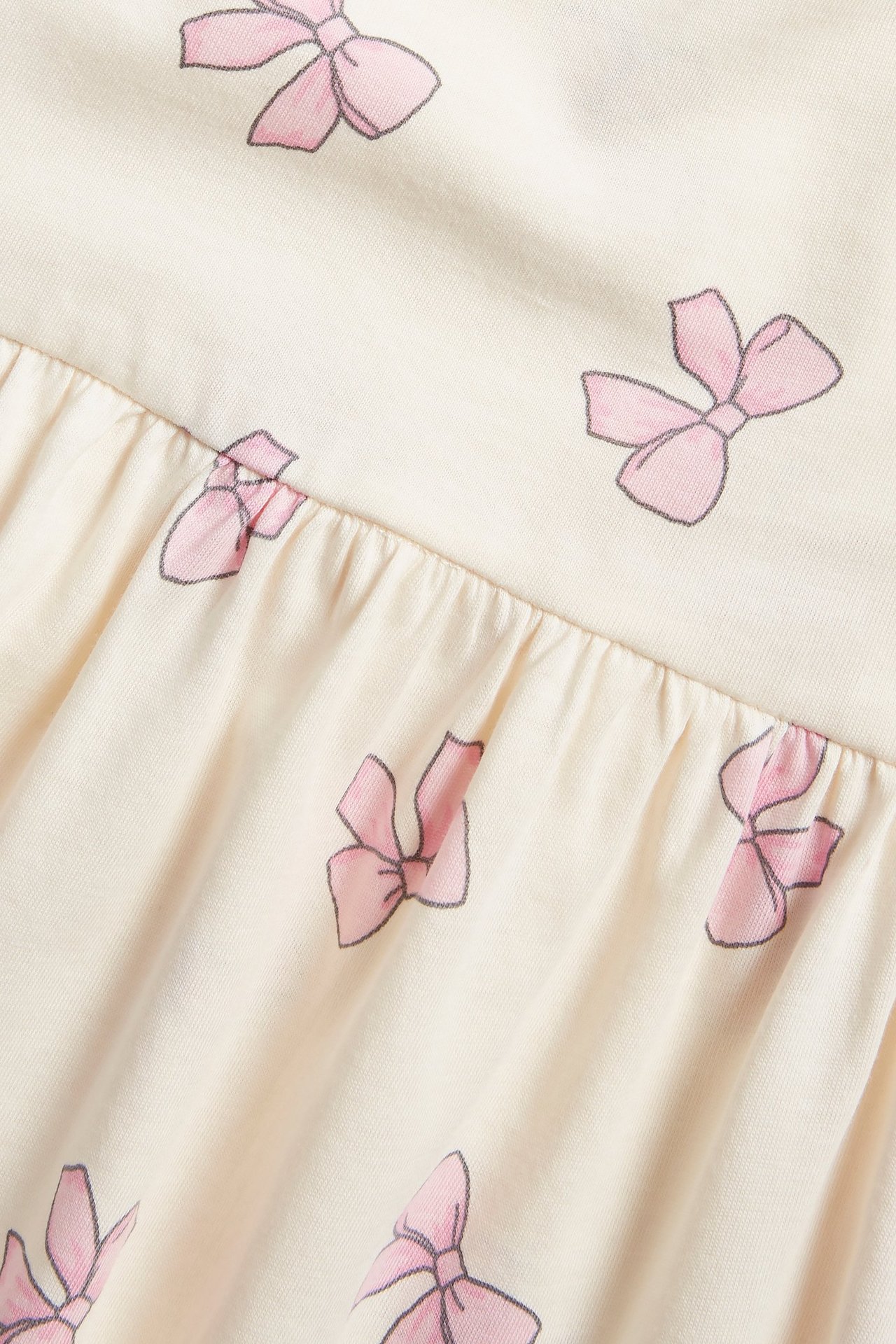 Zbliżenie na sukienkę z bawełnianej dzianiny w kolorze ecru, z różowym wzorem kokardek i marszczonym szwem.