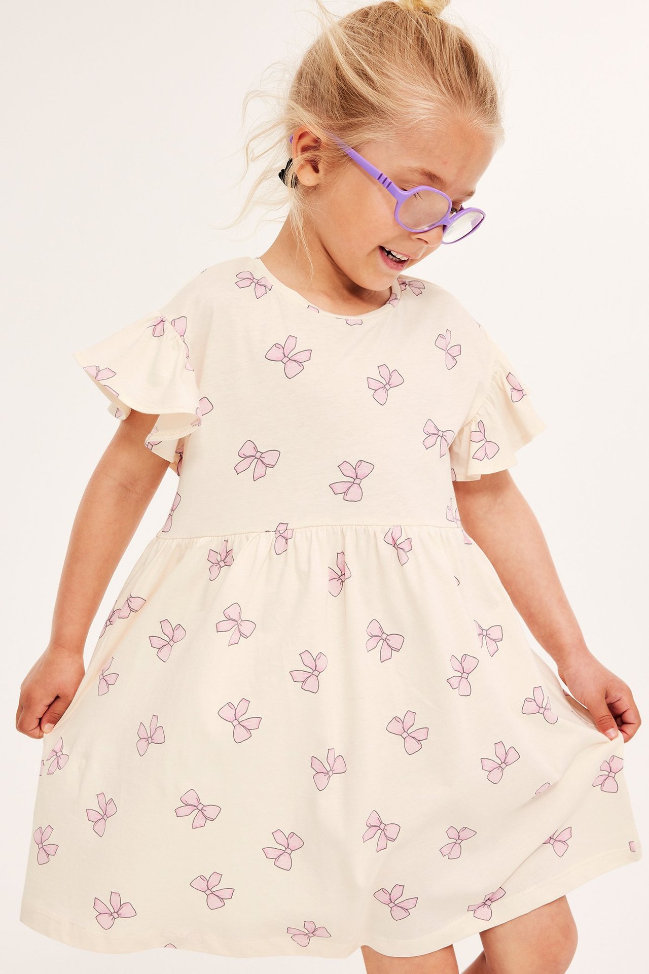 Widok z przodu dziecka w sukience z bawełnianej dzianiny w kolorze ecru z różowymi kokardkami i fioletowymi okularami.
